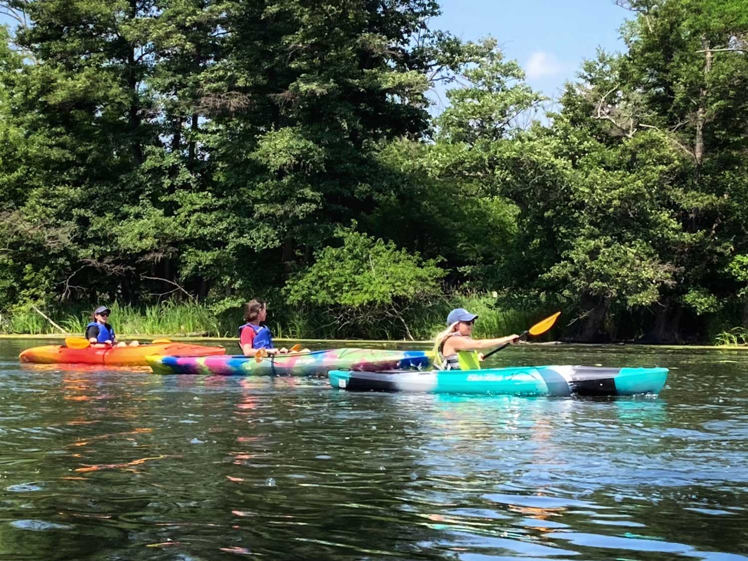 Three people sitting in colorful kayaks on the water. One of the people is holding an oar above the water.
