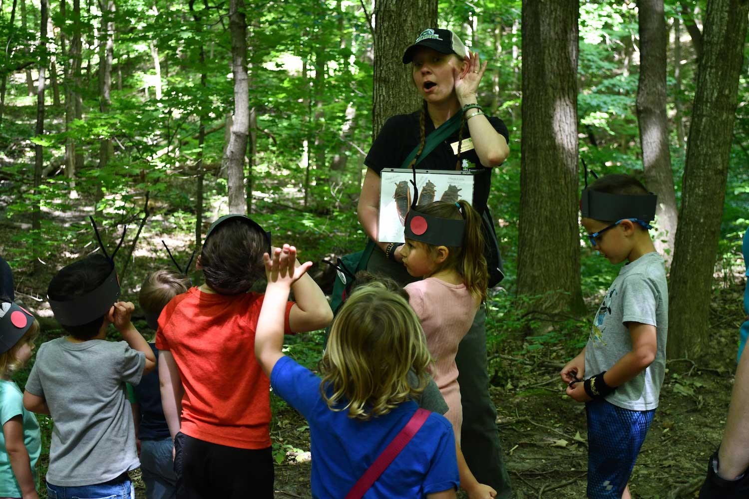 An adult with a hand to their ear standing in the woods in front of a group of six young kids. Some of the kids also have their hands to their ears and one has their arm raised.