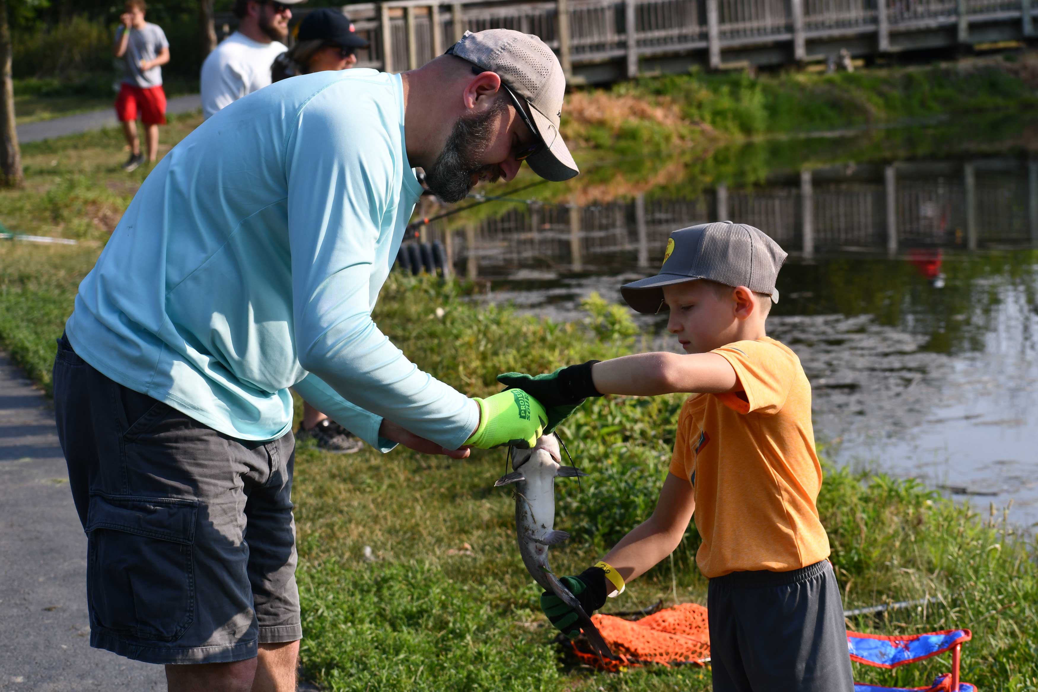 An adult helping a child hold a large fish while standing in a grassy area between a trail and the water.