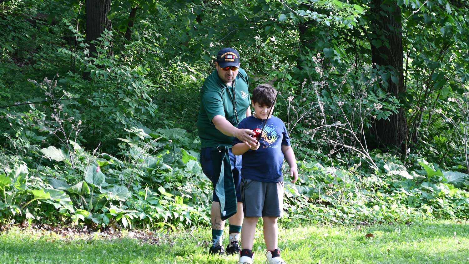 An adult standing behind a child holding a fishing pole instructing them how to use it.