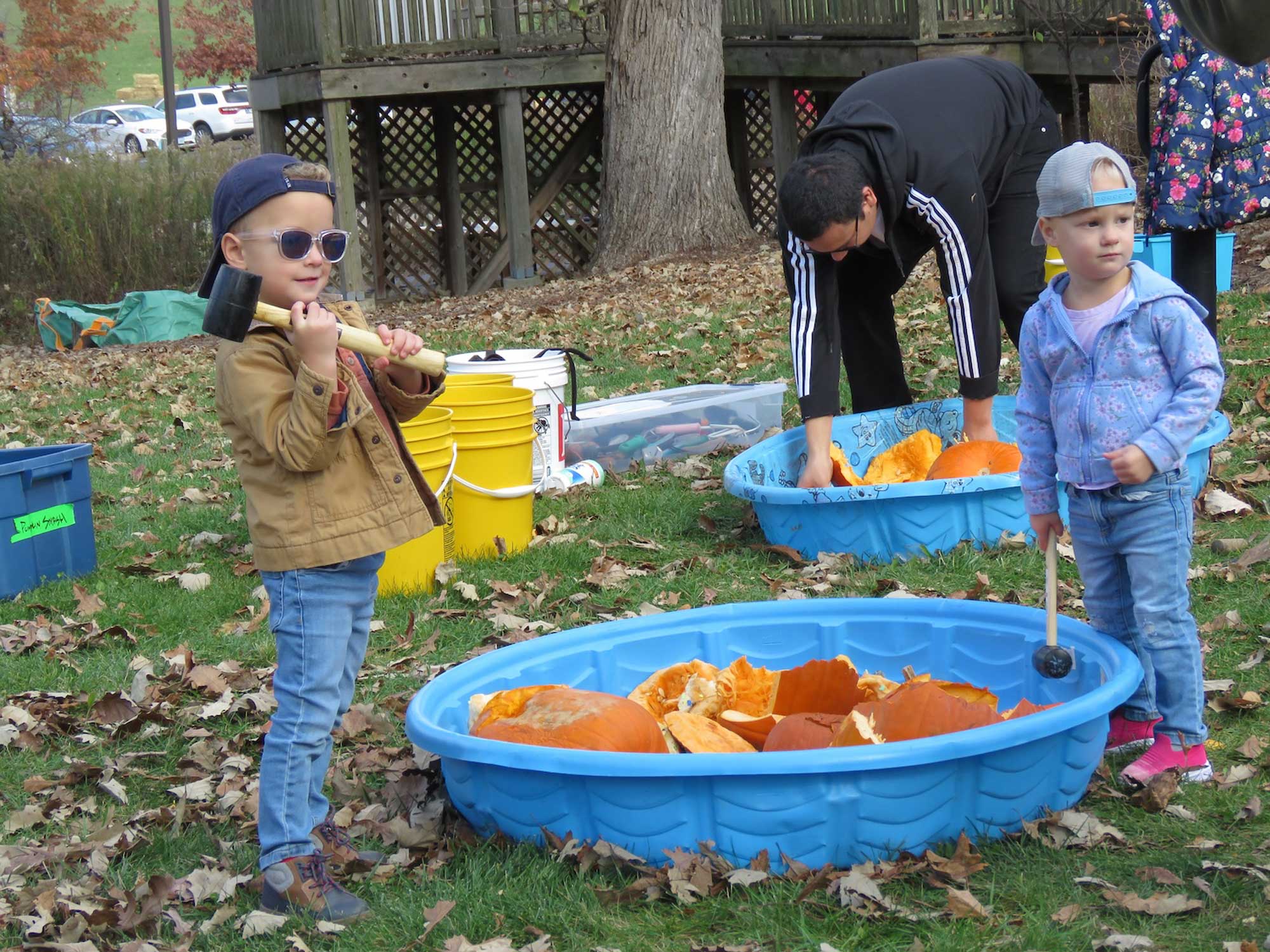 Two kids holding mallets while standing outside a kiddie pool filled with pumpkins. 