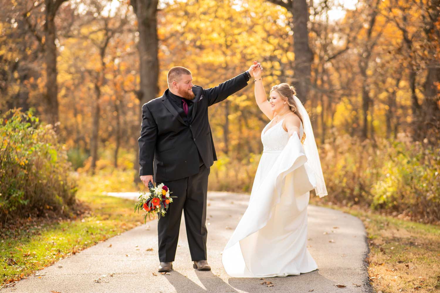 Newly married couple standing on paved trail with trees in the background.