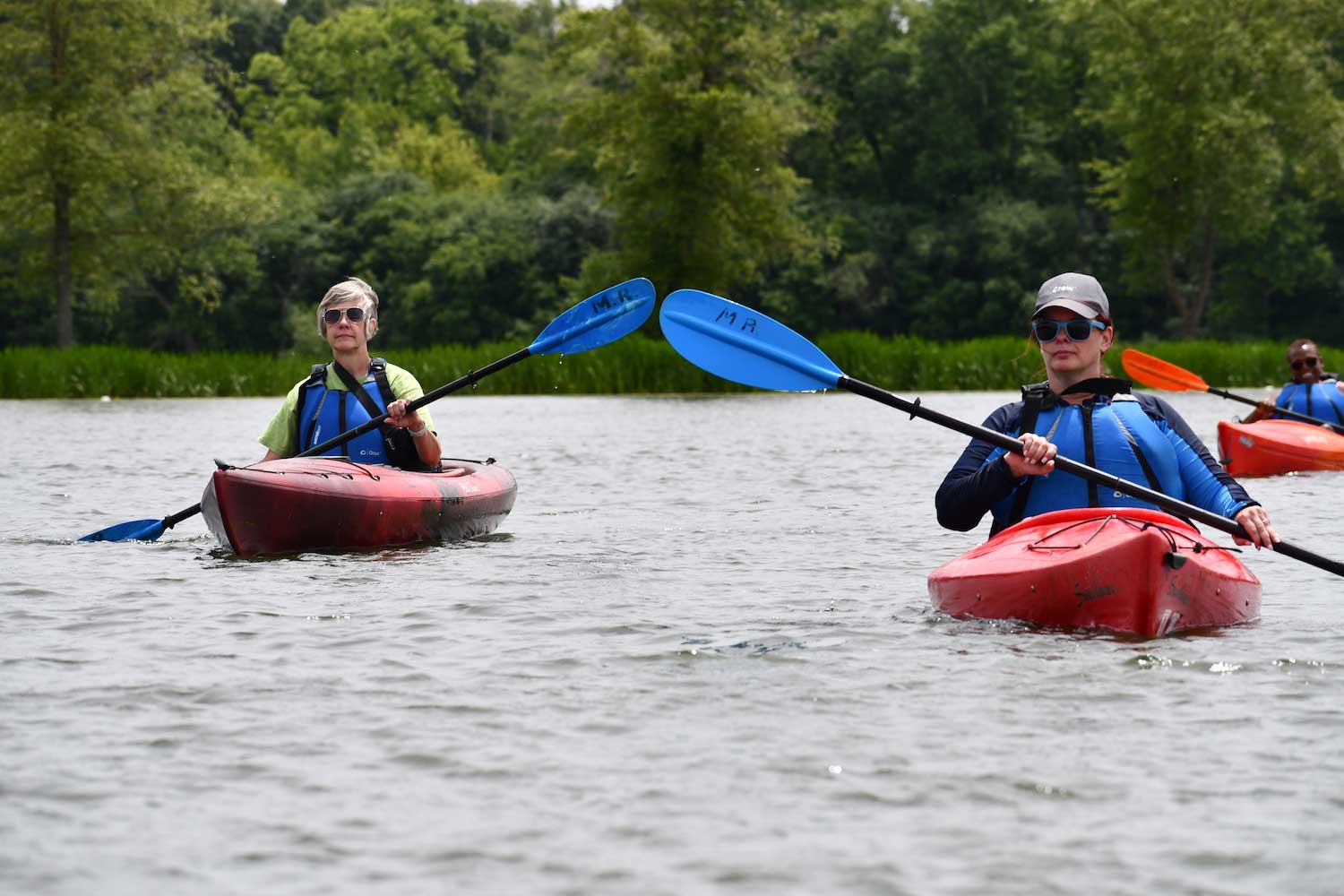Three red kayaks on water, with people inside holding colorful paddles up over the water. 