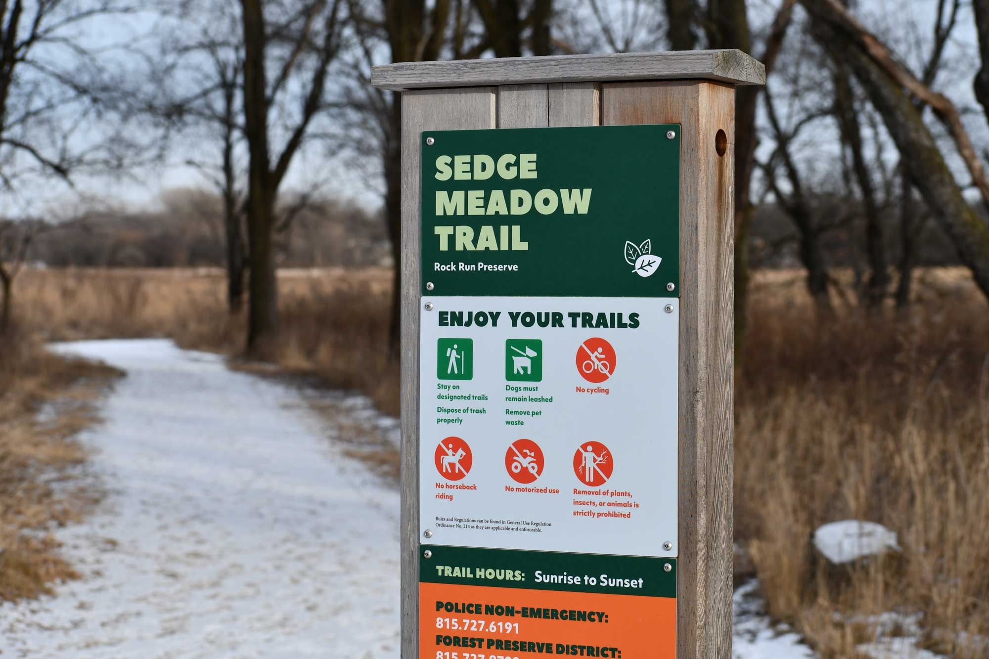 Trail sign with vegetation and a snow covered trail in the background.