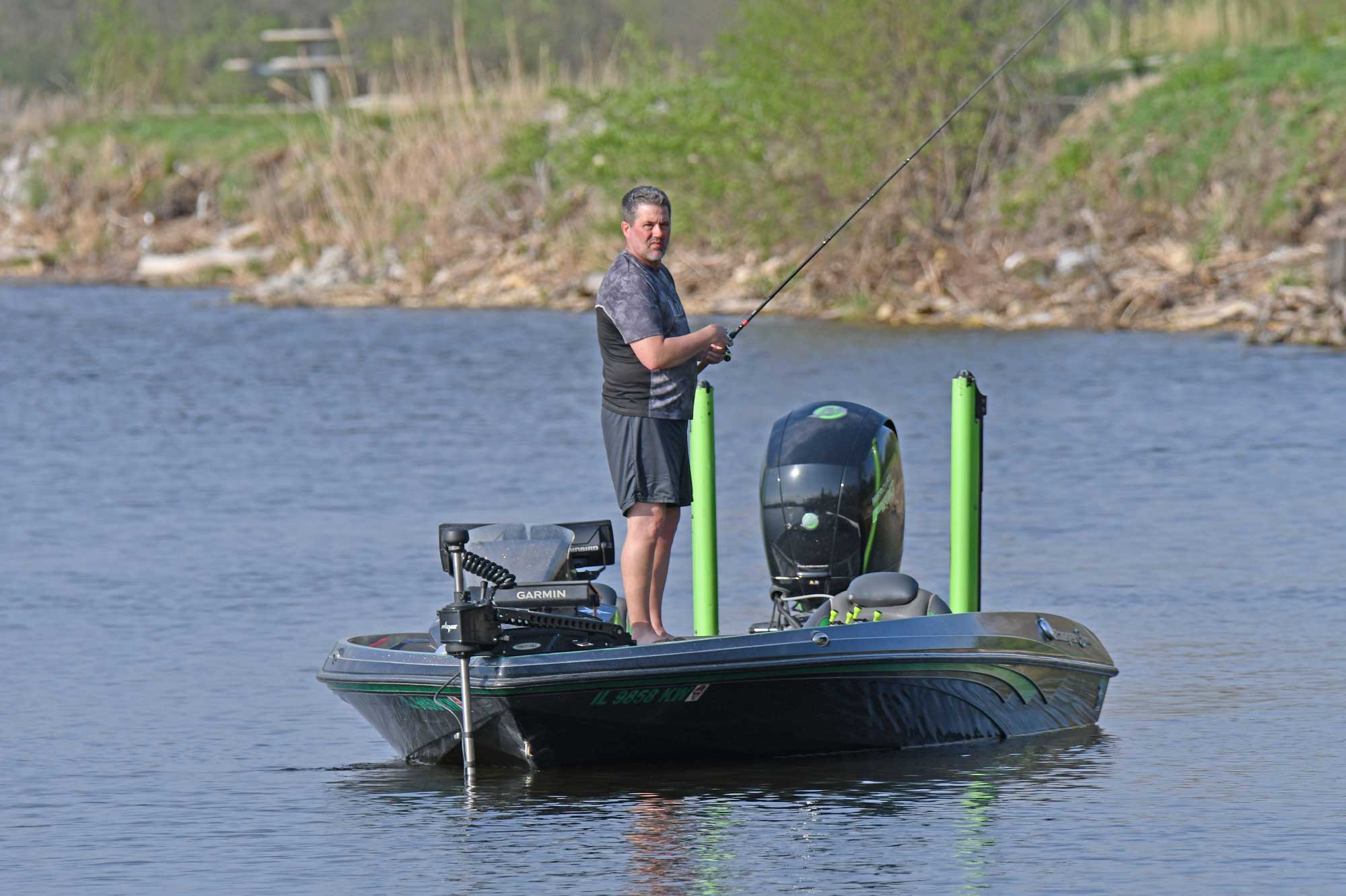 Person standing up in a boat while fishing with vegetation in the background.