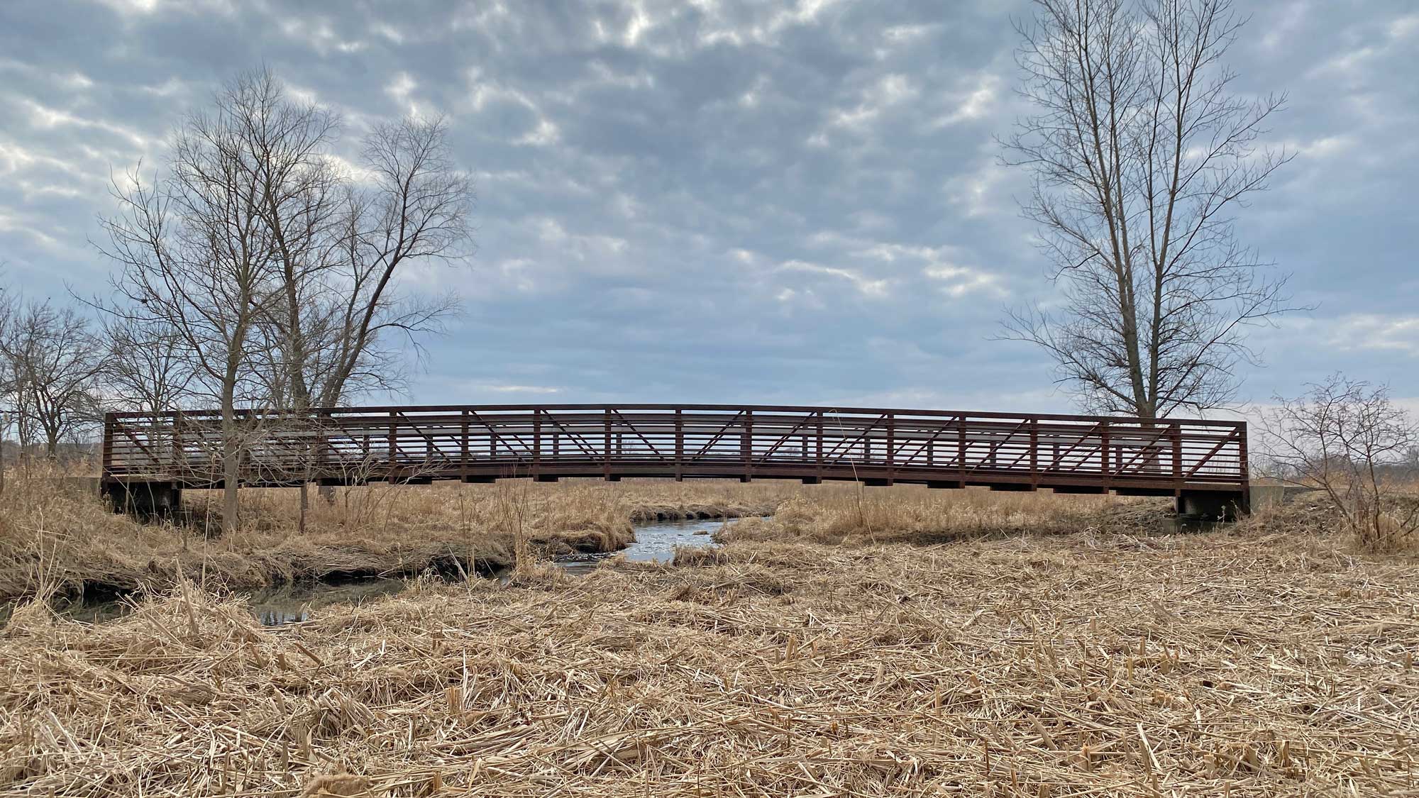 A scene showing a prairie, with a long, brown metal bridge spanning a creek. 