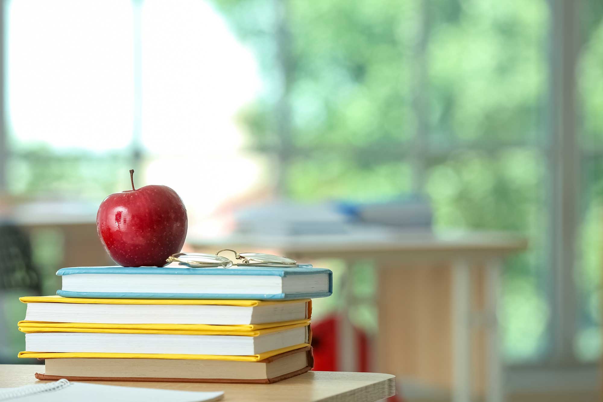 A red apple sitting on top of a stack of books in a classroom setting with trees visible outside the window.
