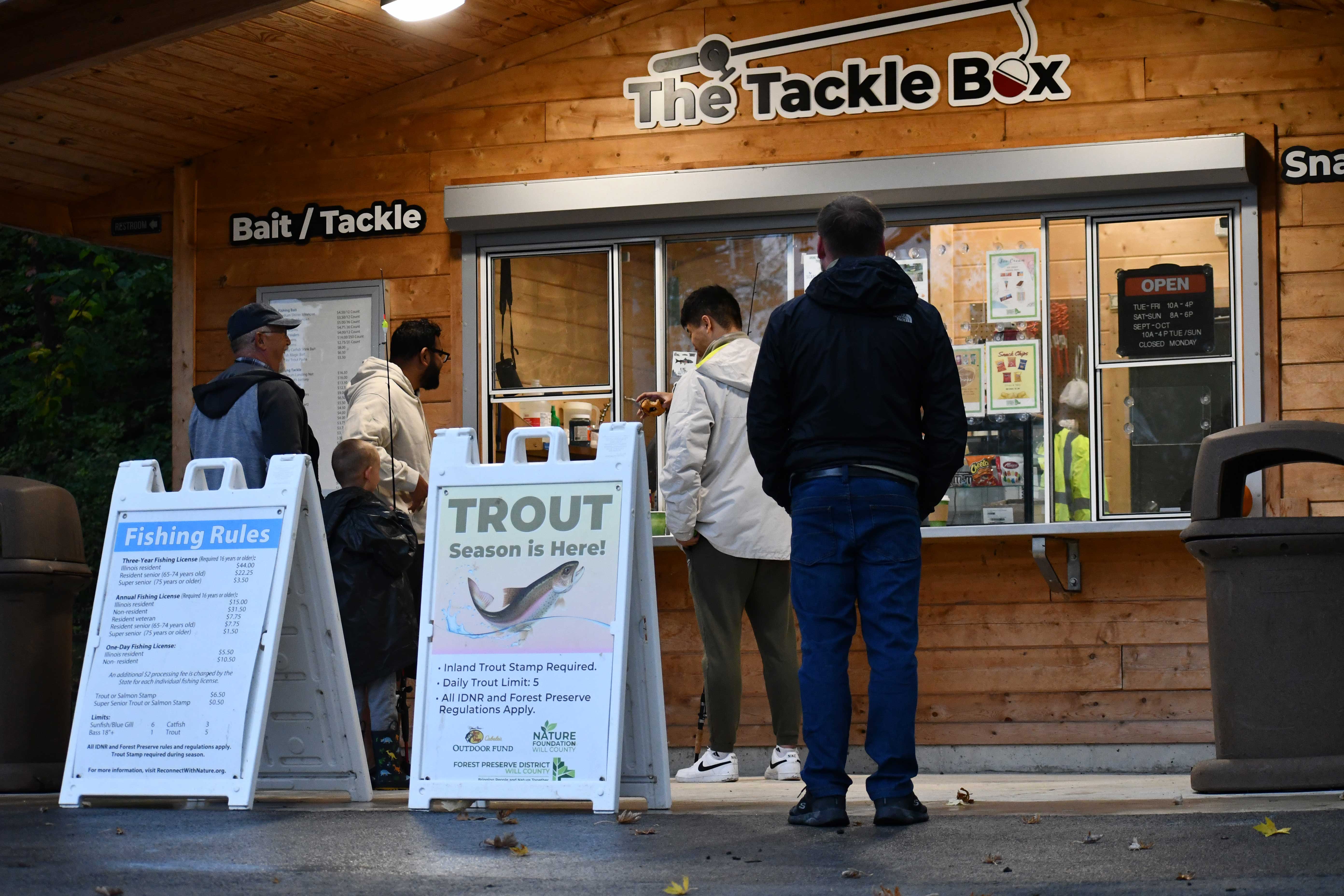 Several people stand in line outside “The Tackle Box,” a wooden bait and tackle shop. Signs in front advertise fishing rules and announce that trout season is open.