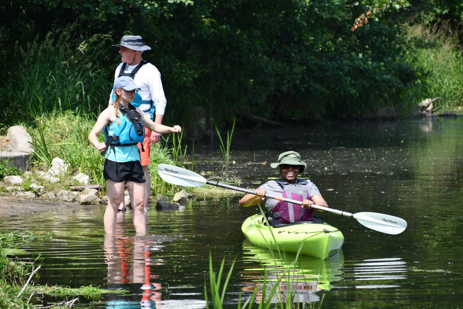 A person standing in shallow water next to a person in a kayak in the water. Another person wearing a life jacket is standing next to the shoreline in the background.