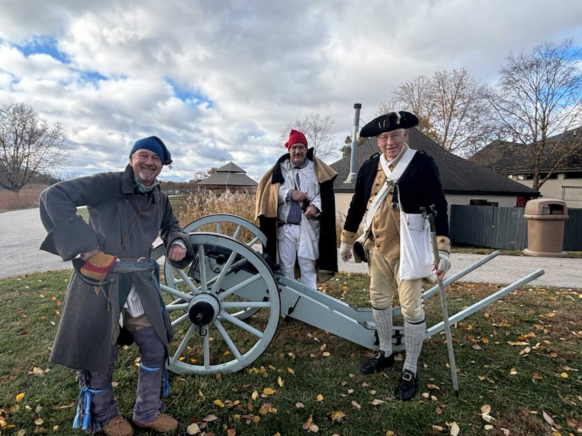 Three people in 18-century clothing standing around a cannon in the grass.