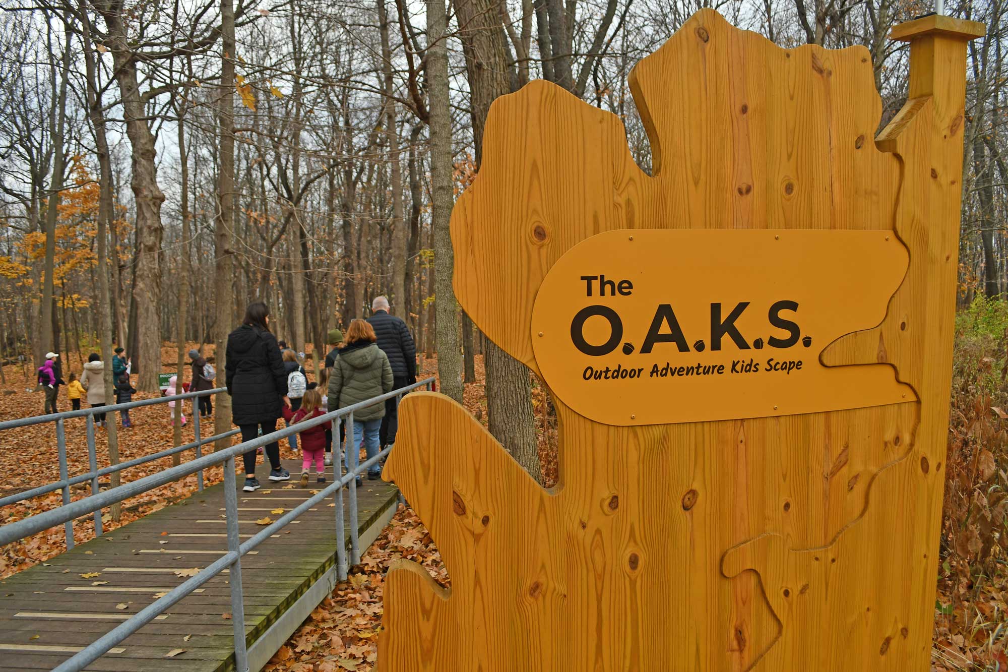 A group of kids and adults walking down a path into a woodland. In the foreground is a sign stating Outdoor Adventure Kids Scape.