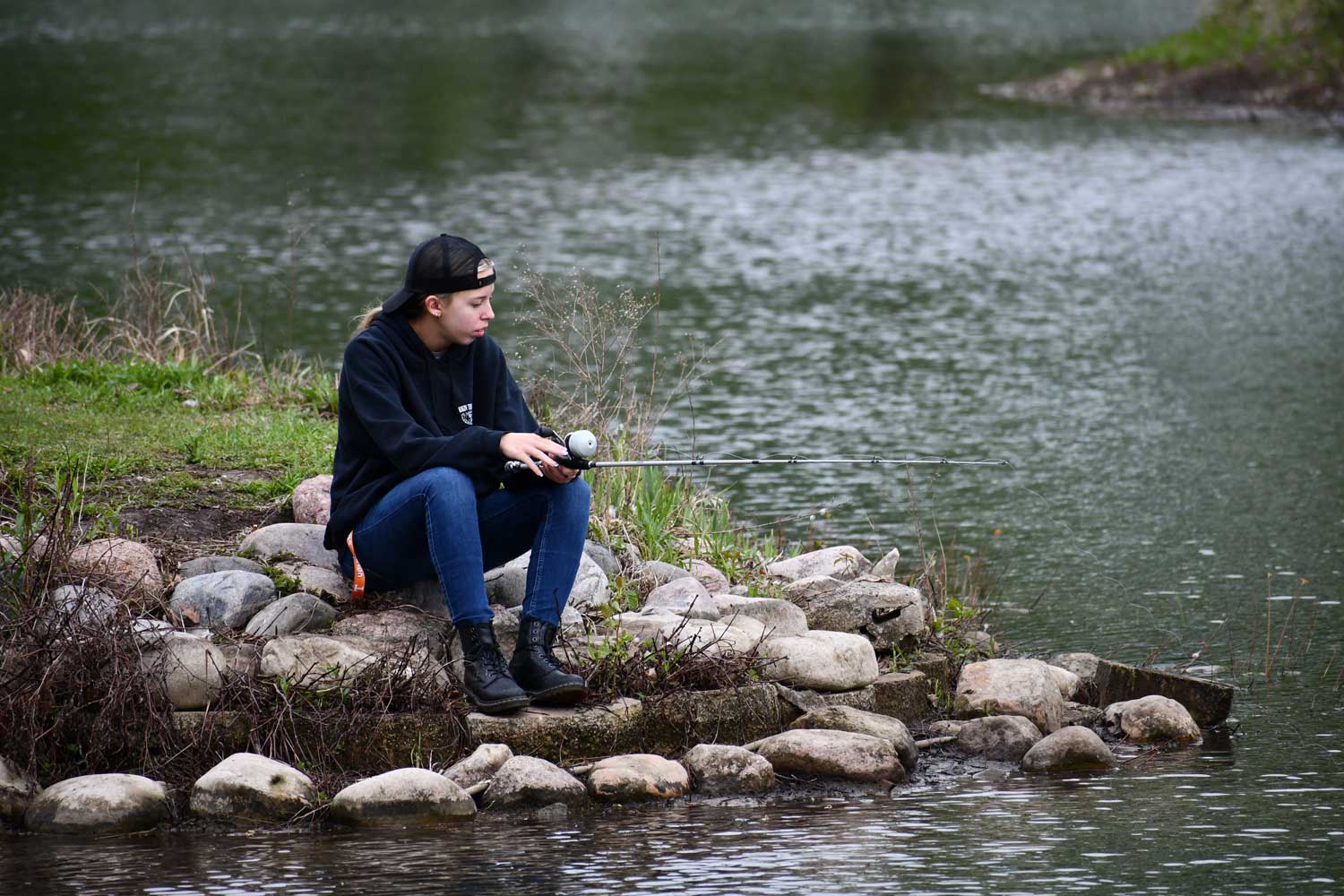 A person sitting on rocks along a shoreline while holding a fishing pole with a line in the water. 