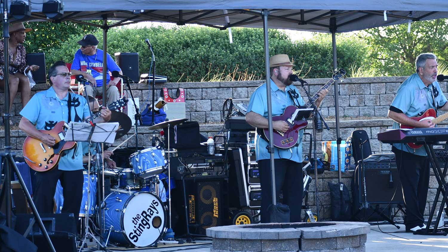 Three guitarists playing their instruments under a tent with drums and other band equipment around them. 