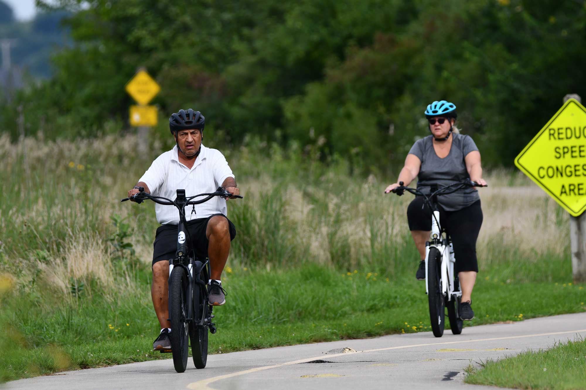 Two people biking on a paved trail with vegetation and trees in the background.