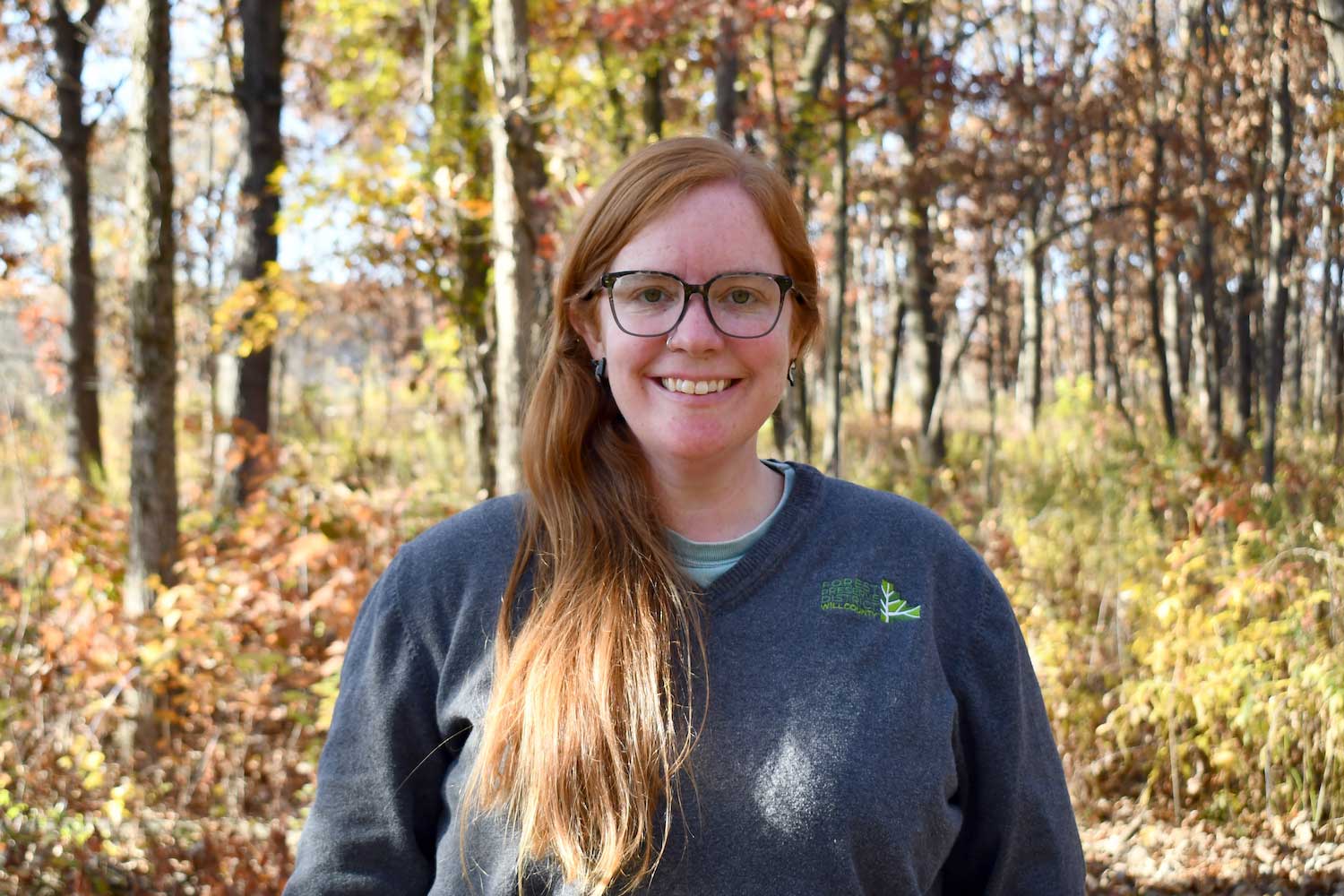 A person standing in a forest with the trees in the background partially full of yellow, orange and brown leaves.