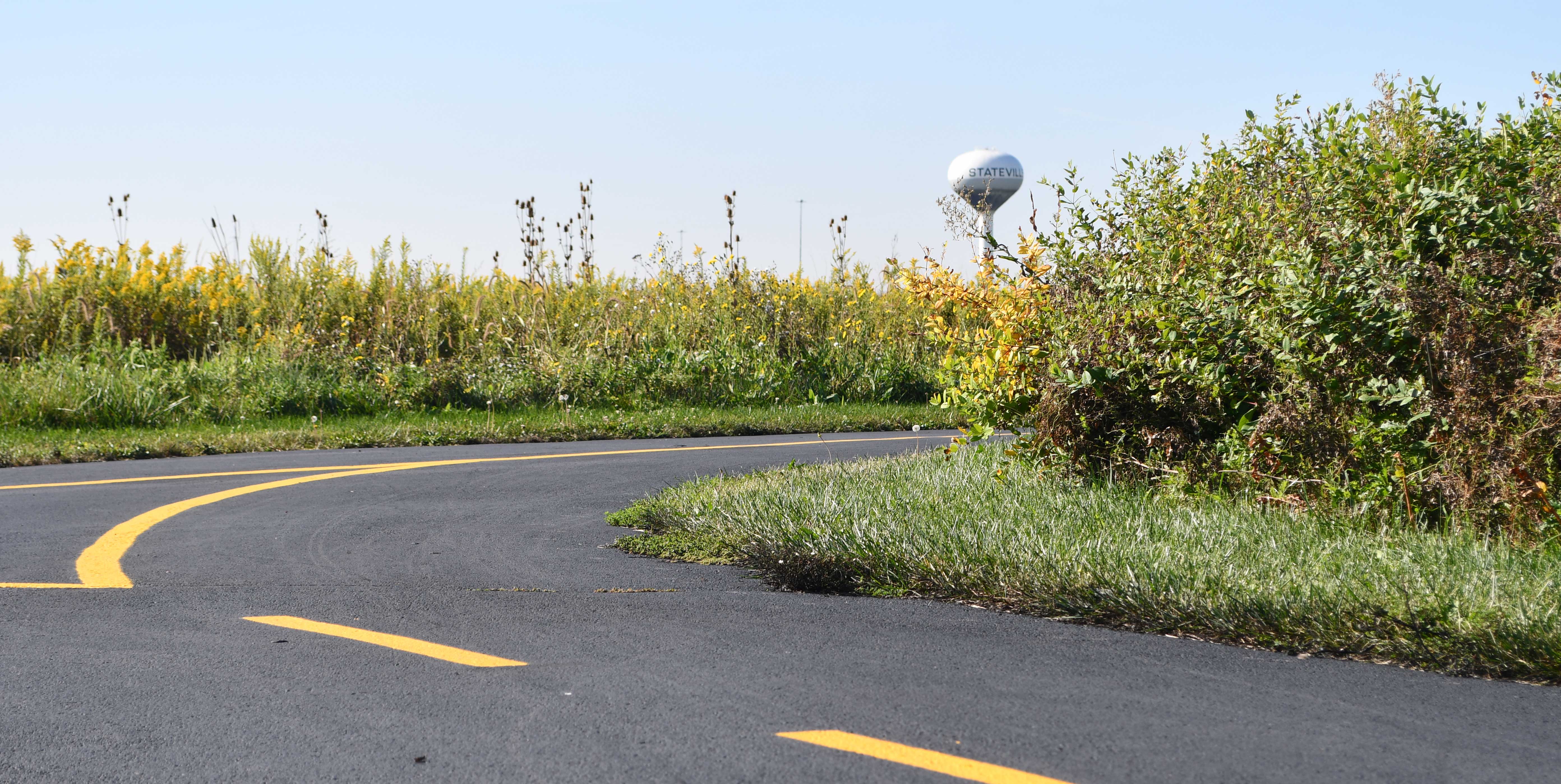 Curved asphalt trail lined with yellow wildflowers and dense greenery at Prairie Bluff Preserve, with a distant view of the Stateville water tower under a clear blue sky.