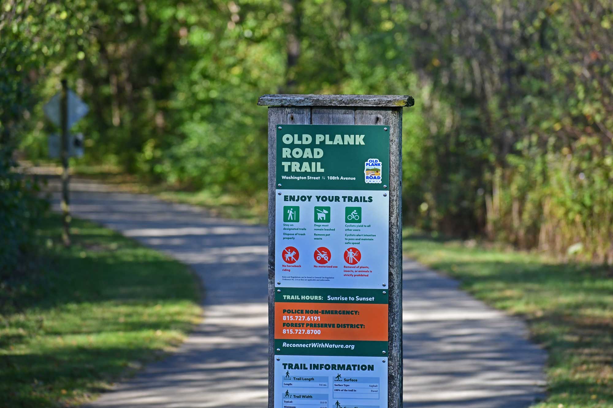 Trail sign with paved trail and trees in the background.