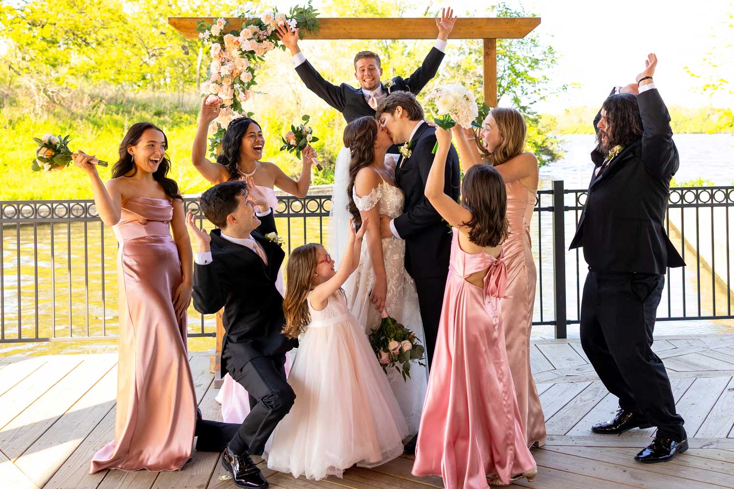 Wedding party surrounding newly married couple as they share a kiss.