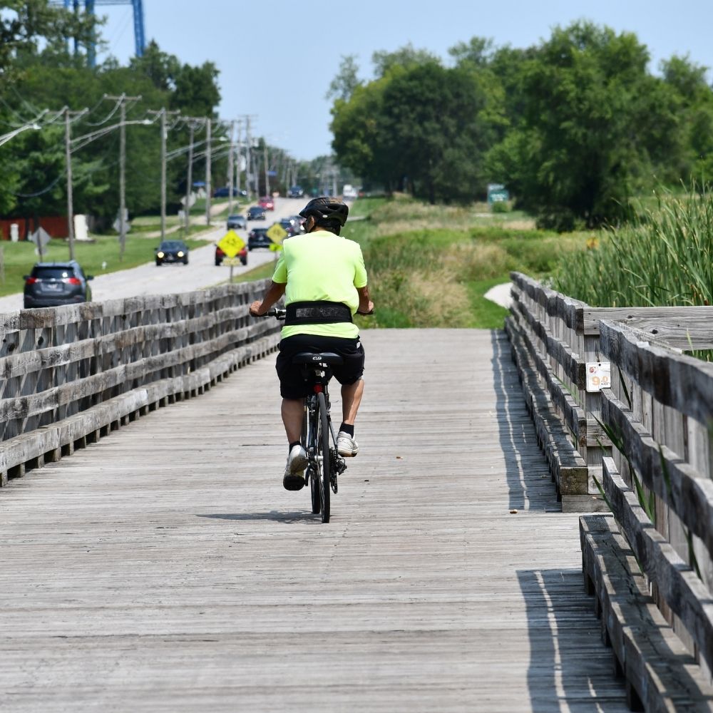 A cyclist wearing a bright neon shirt and helmet rides across a long wooden boardwalk trail on a sunny day.
