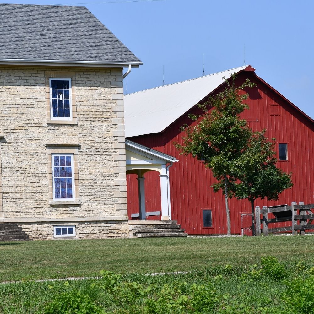 A historic farmstead featuring a light-colored stone farmhouse beside a large red barn under a clear blue sky.