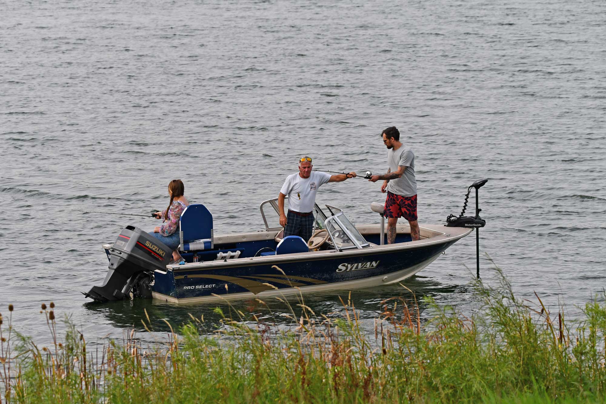 Three people in a boat fishing near some tall grasses.