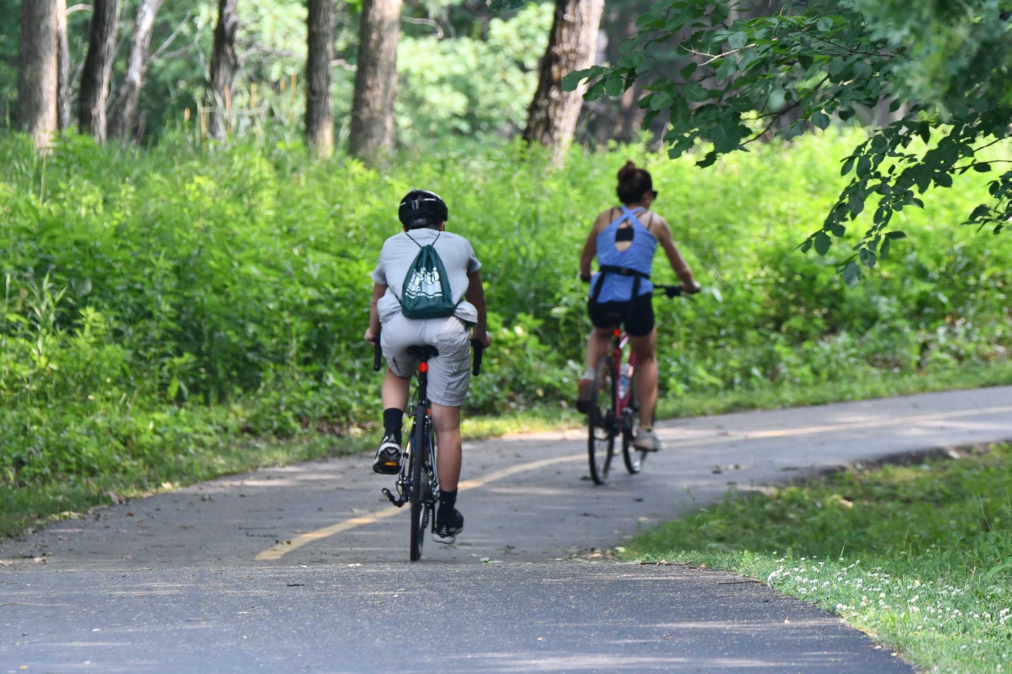 People riding bikes on a paved trail with vegetation and trees in the background.