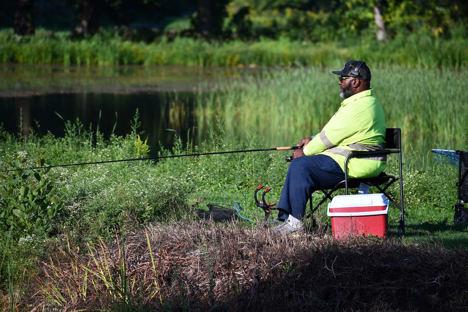 A person sitting in a yard chair with a fishing pole cast in a lake. A red cooler is next to the chair.