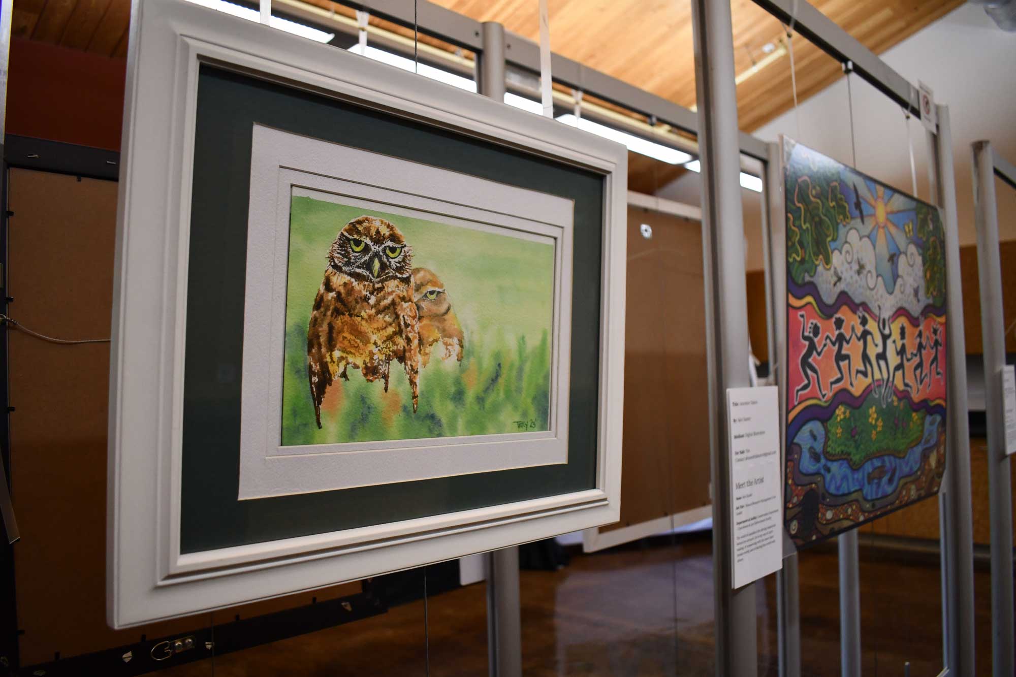 A framed painting of an owl displayed on a wall with another painting visible in the background.