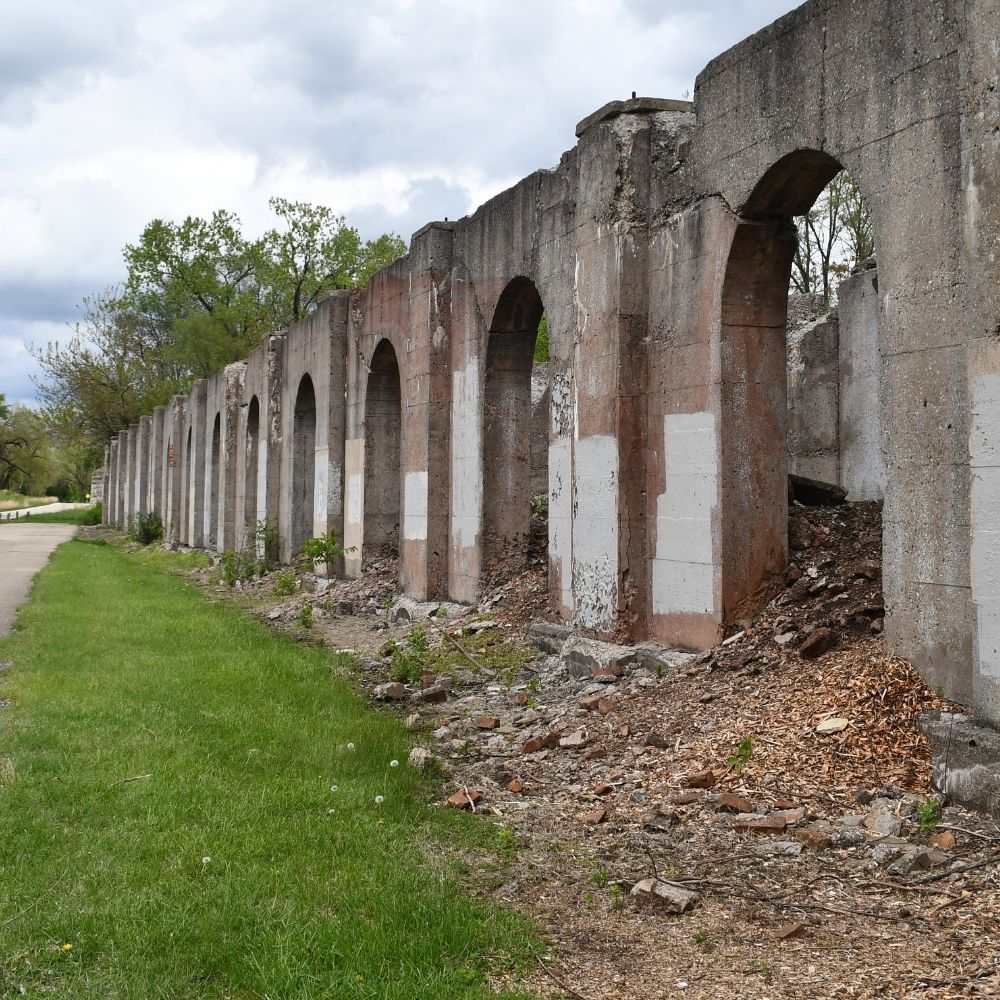 A long row of weathered concrete arches from a ruined industrial structure stretches into the distance beside a grassy path under cloudy skies.
