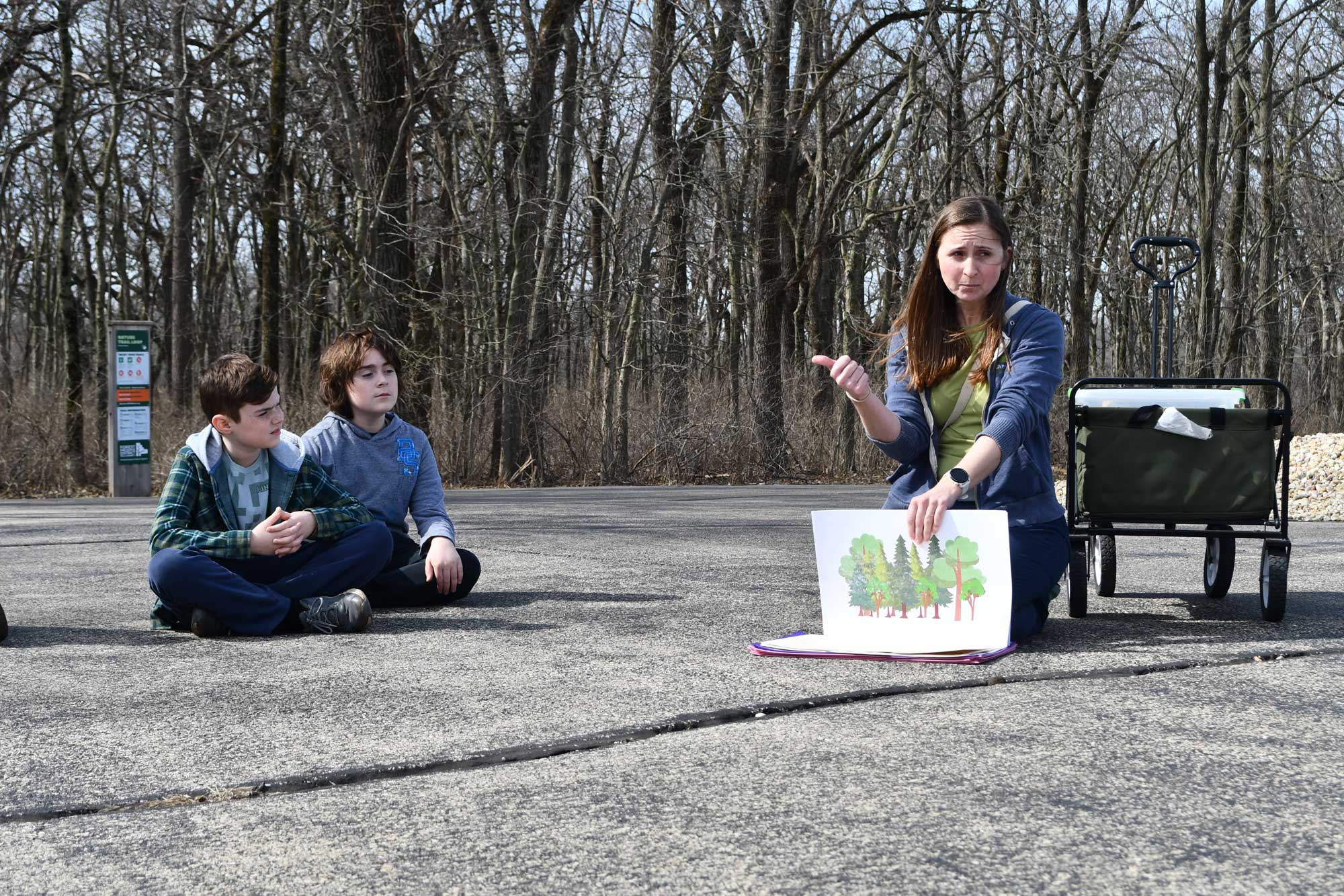 An adult and two kinds sitting on the ground. The adult is holding an illustration and gesturing as if explaining. 