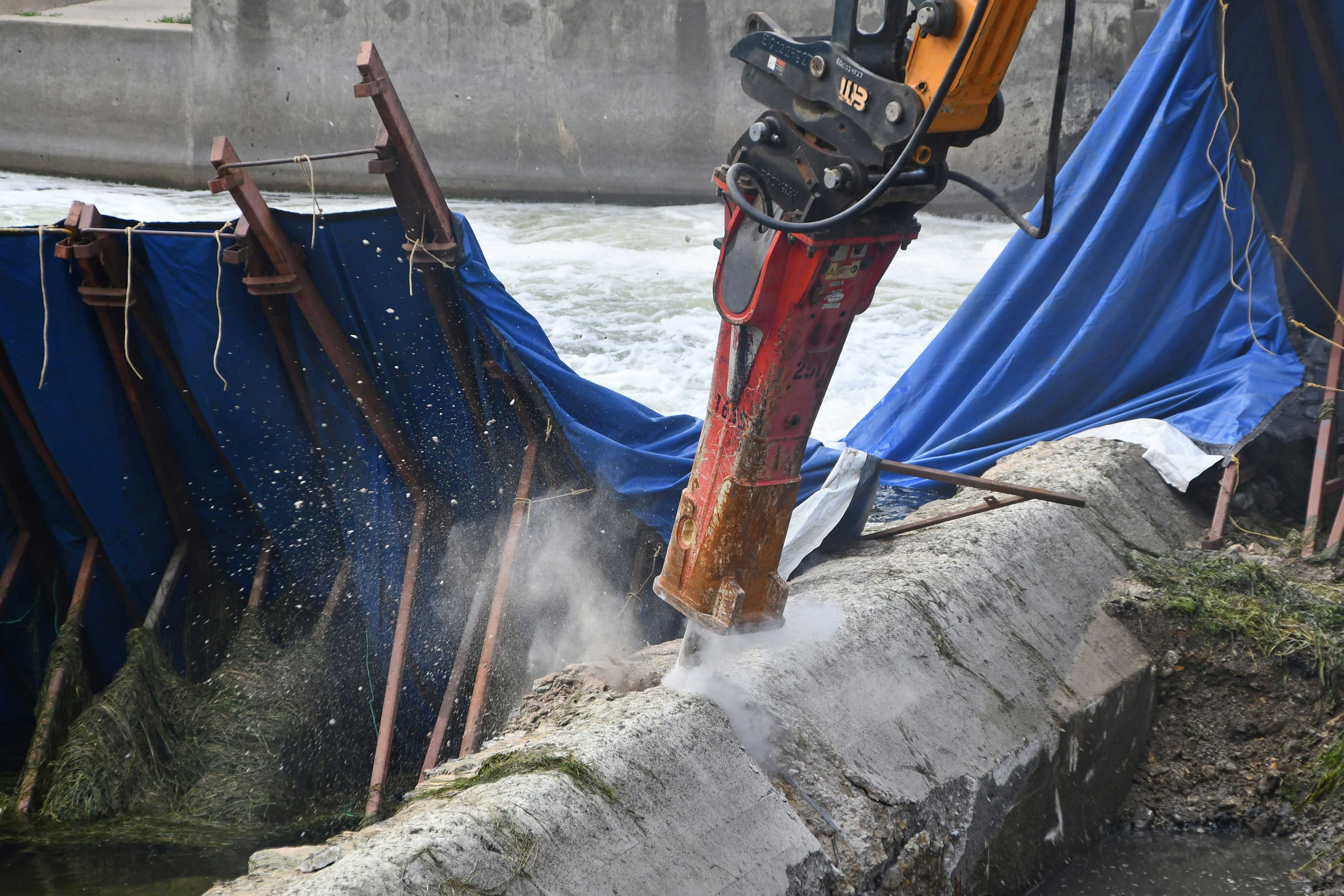 A hydraulic breaker on the end of an excavator chipping away at the dam.