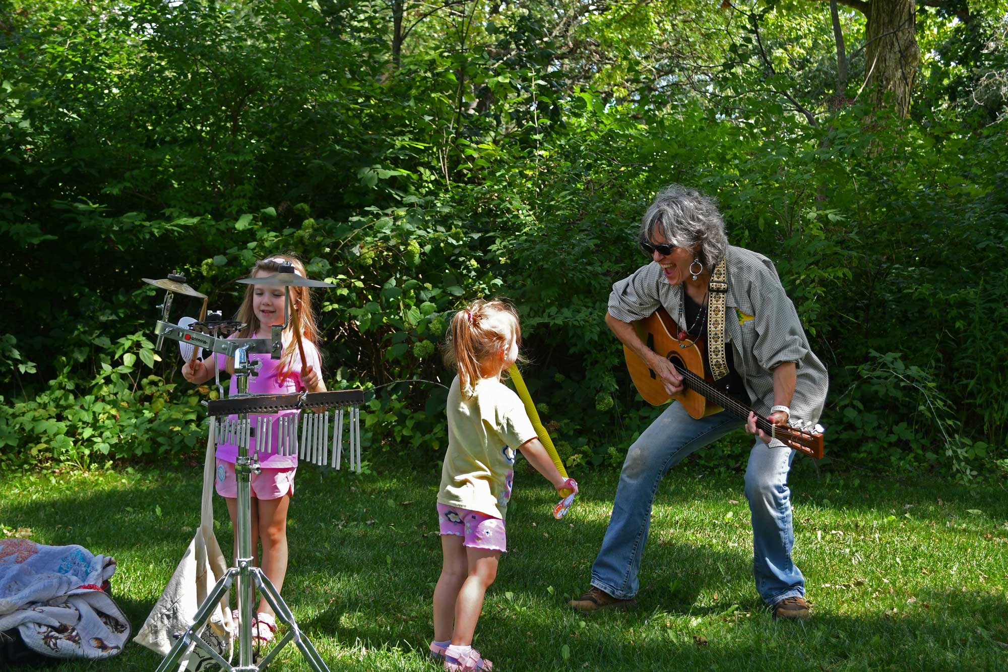 An adult playing guitar on the grass while two children play instruments.