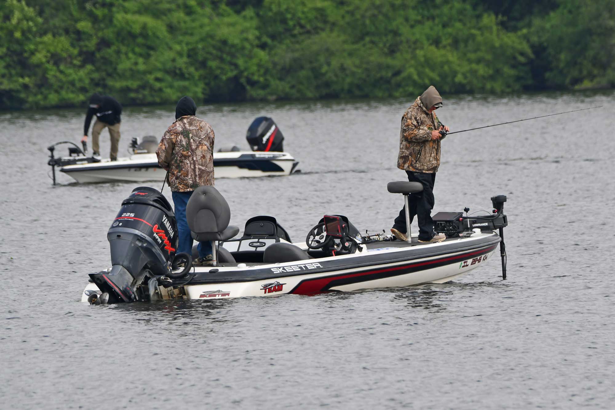 People in a boat while fishing with another boat and trees in the background.