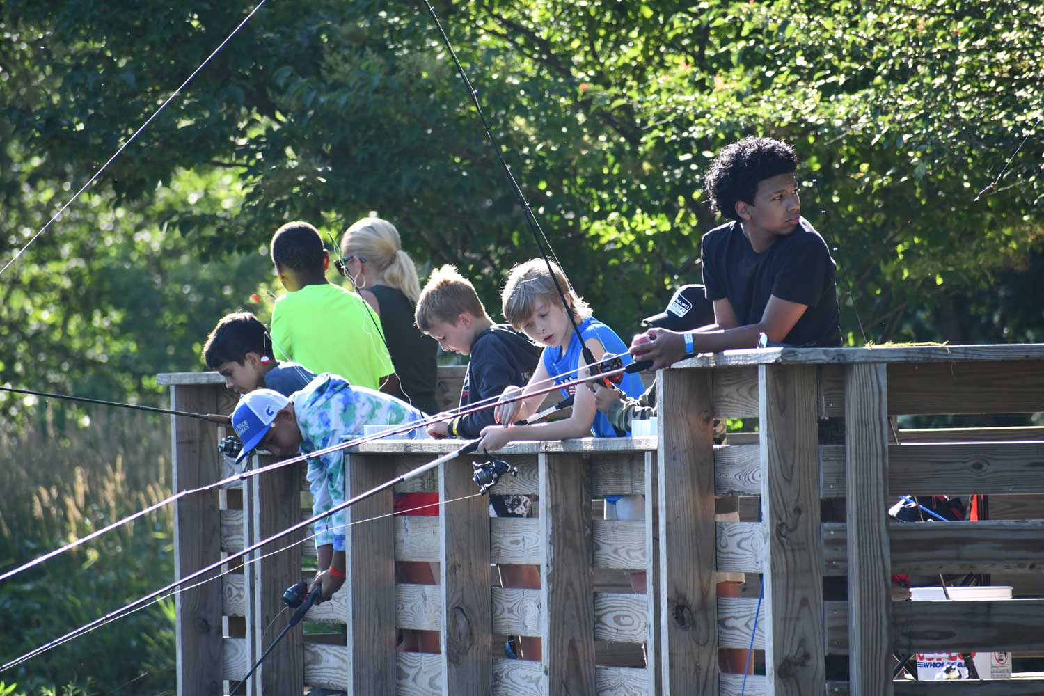 About eight kids lined up against the railing of a wooden pier with fishing poles cast below. 