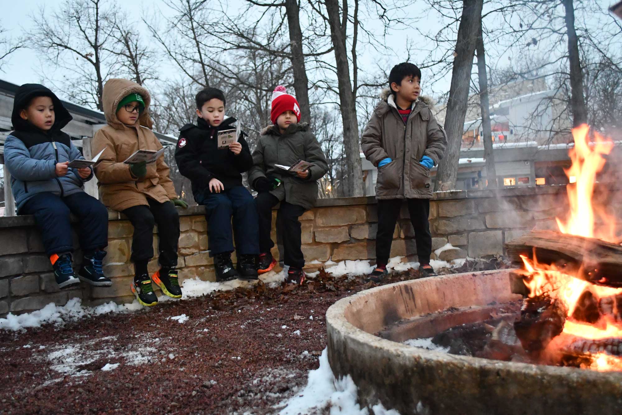 A group of children sitting on a low stone wall around a campfire. The kids are holding caroling books.