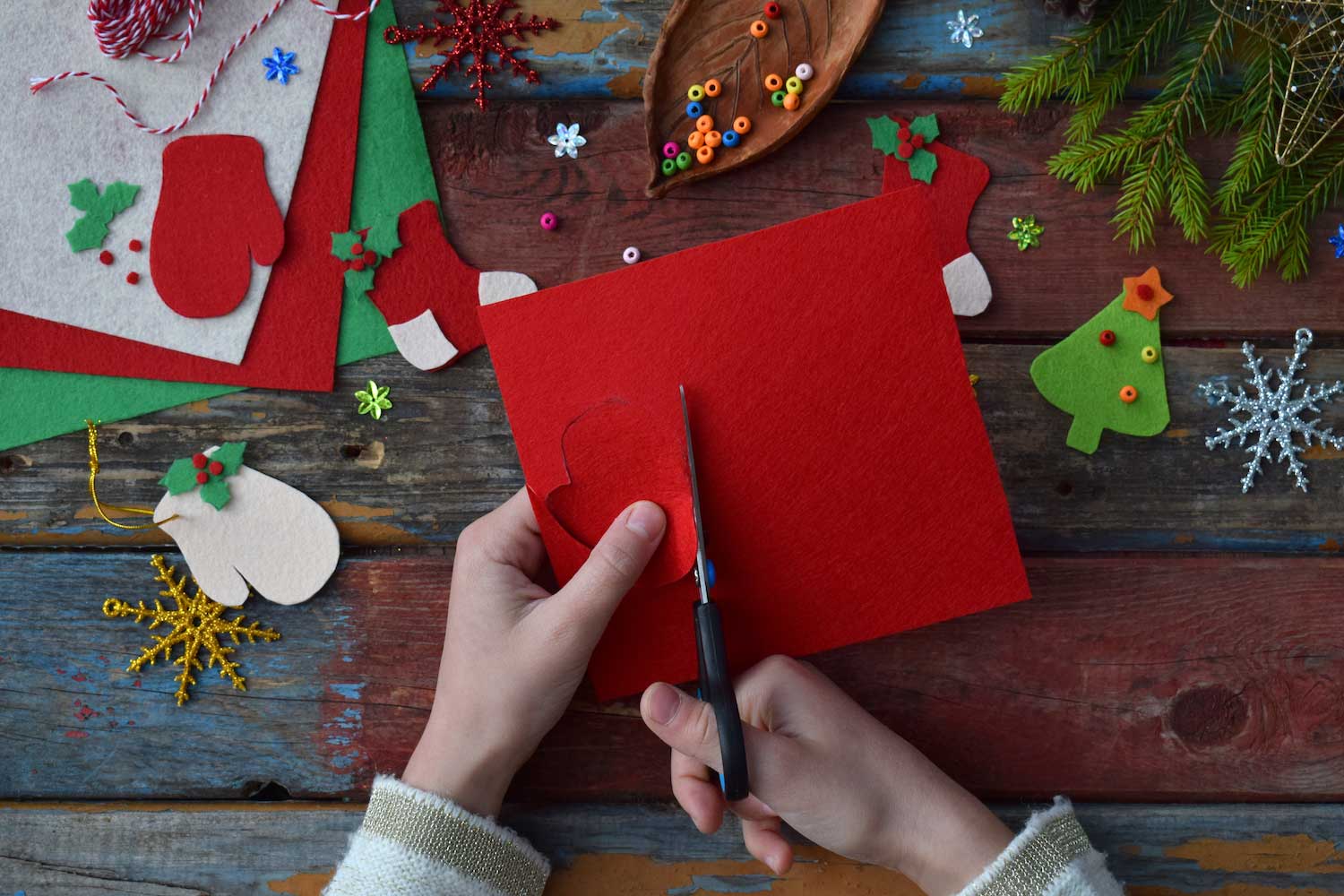 Hands holding scissors cutting red paper over a table with scattered holiday craft supplies on it.