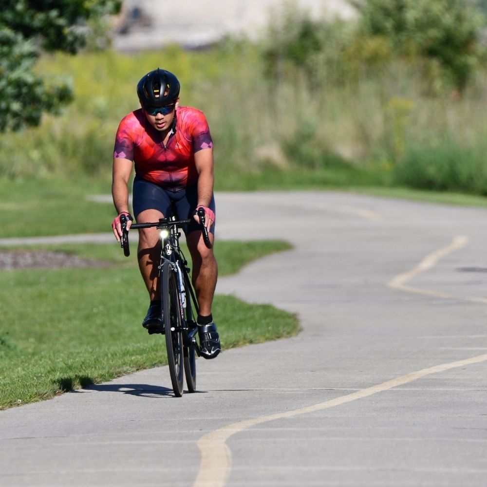 A cyclist rides along a paved trail bordered by green grass and prairie vegetation on a sunny day. 