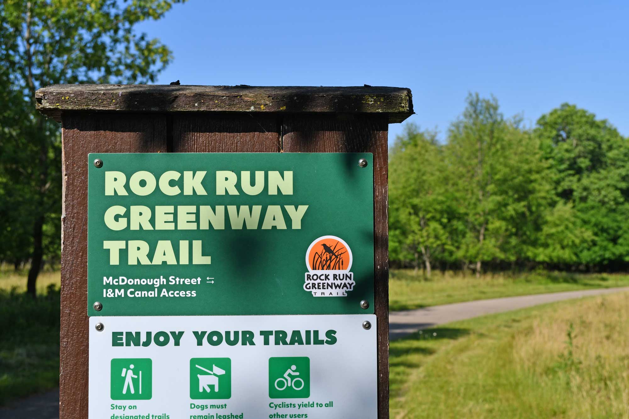 Trail sign with paved trail and trees in the background.