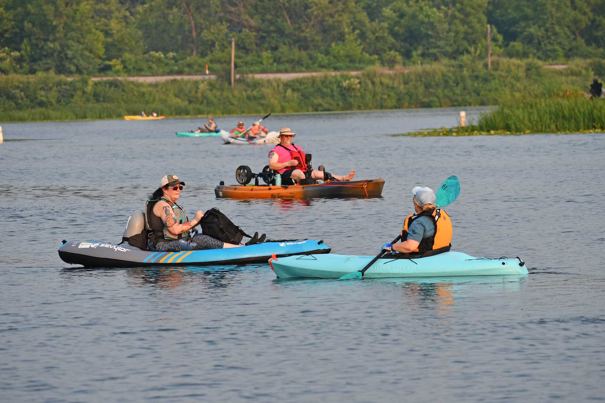 Person kayaking talking to another kayaker with other people boating, water, and trees in the background.
