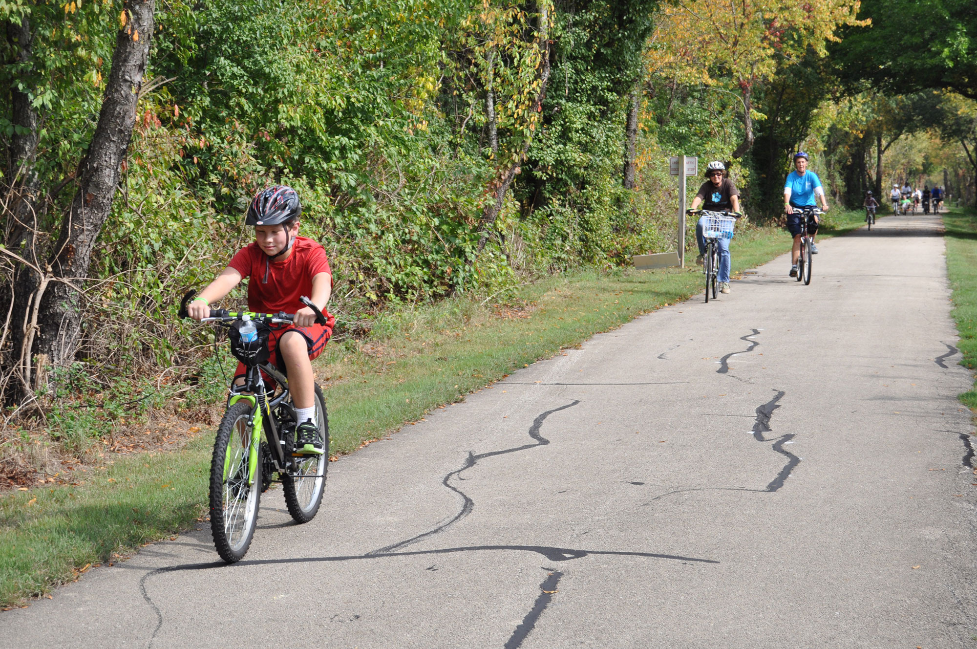 A person riding a bike along a paved trail lined by grass and trees. Two more people on bikes are in the background.