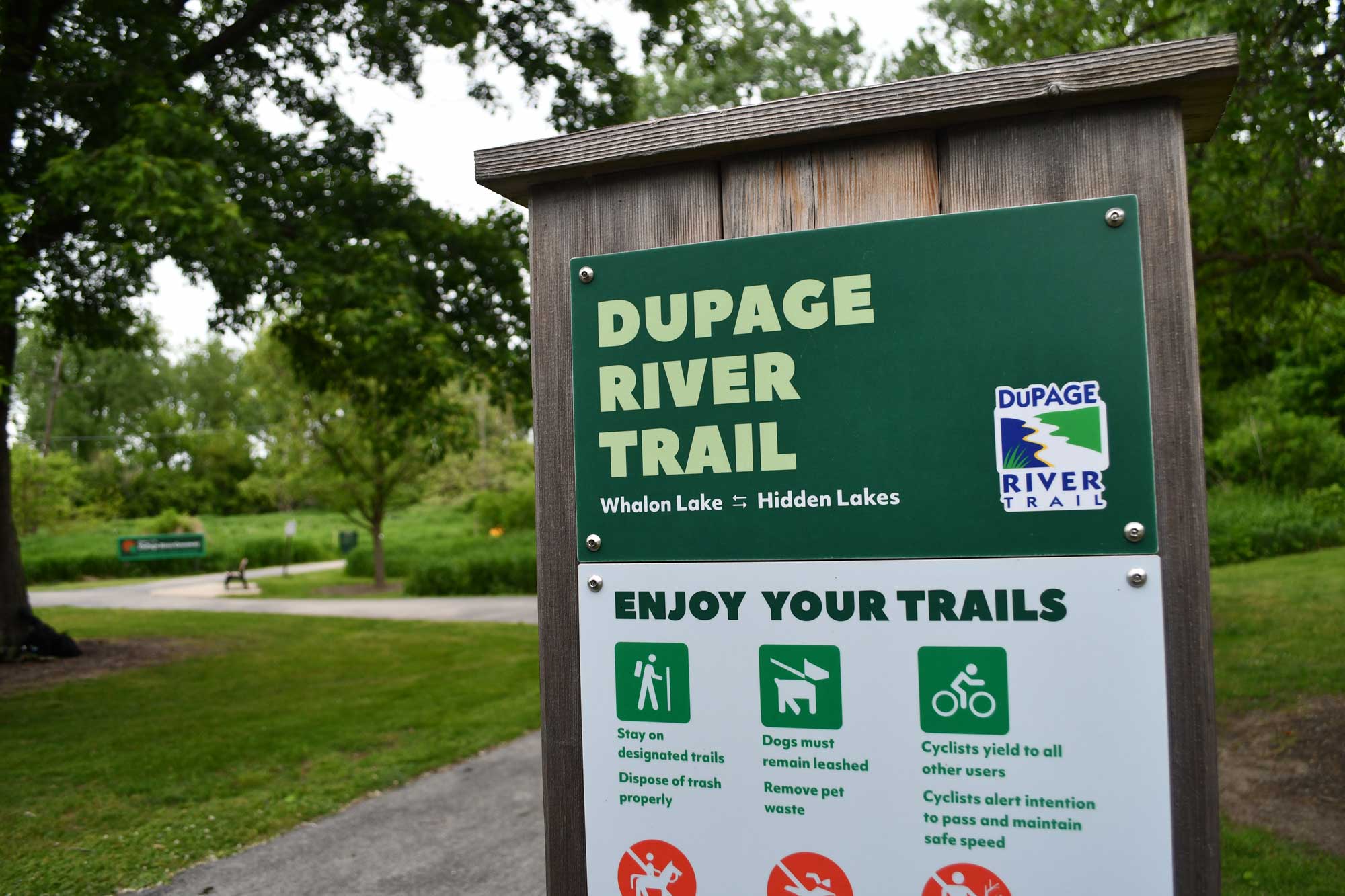 Trail sign with paved trail and trees in the background.