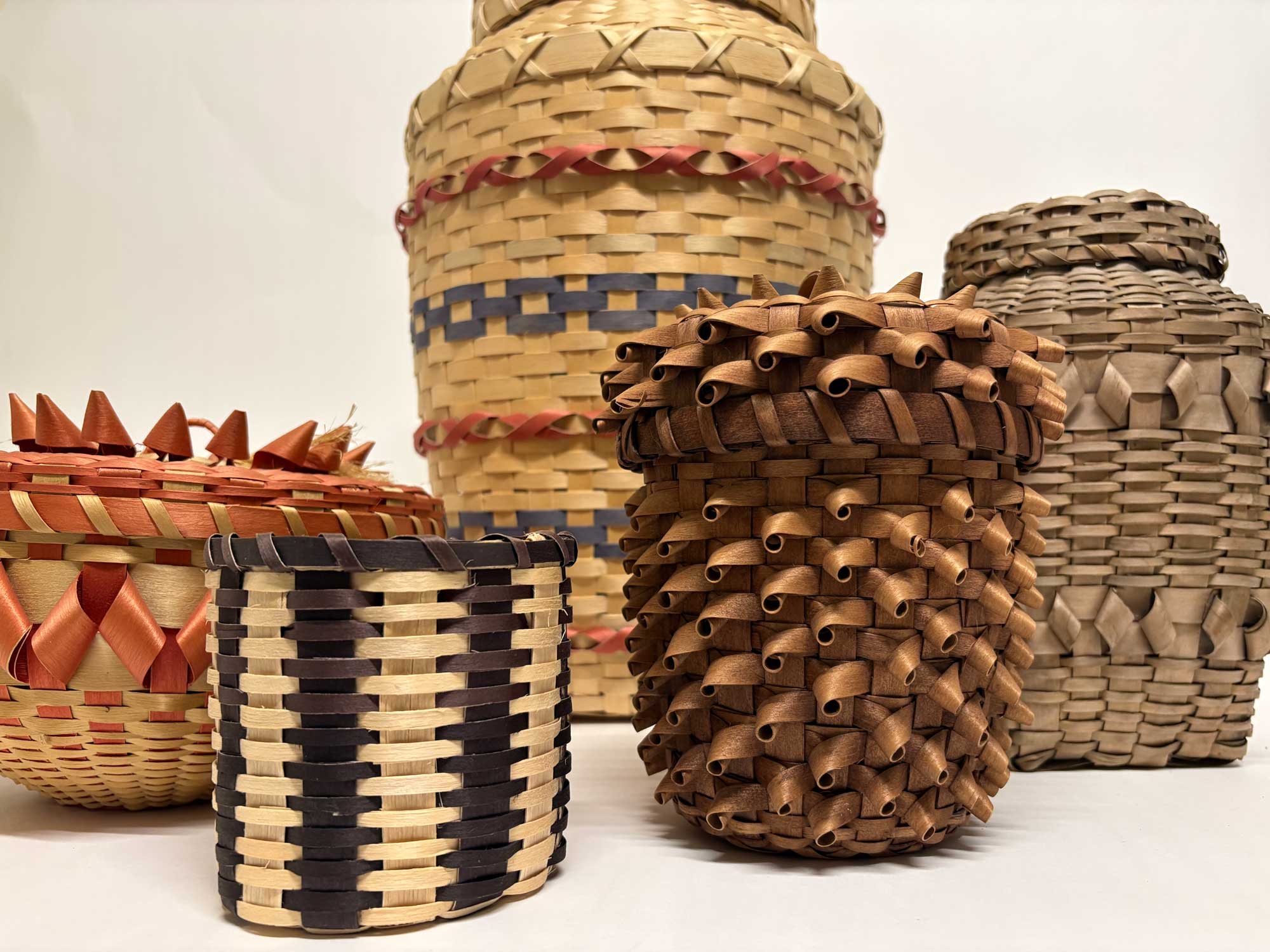 A collection of five woven Potawatomi baskets in shades of brown, red and blue resting on a white surface with a white background.  