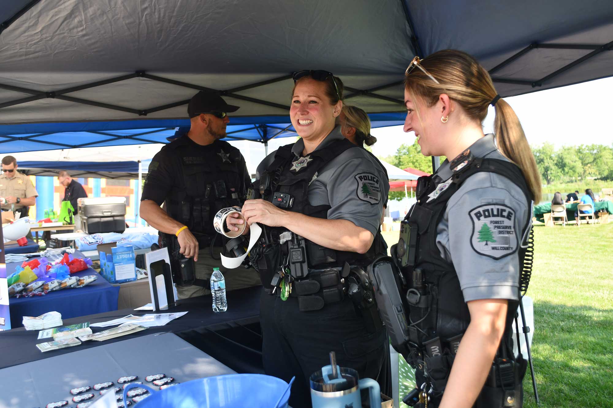 Two police officers in uniform standing at a table under a tent during an outdoor event. One is holding a roll of stickers.