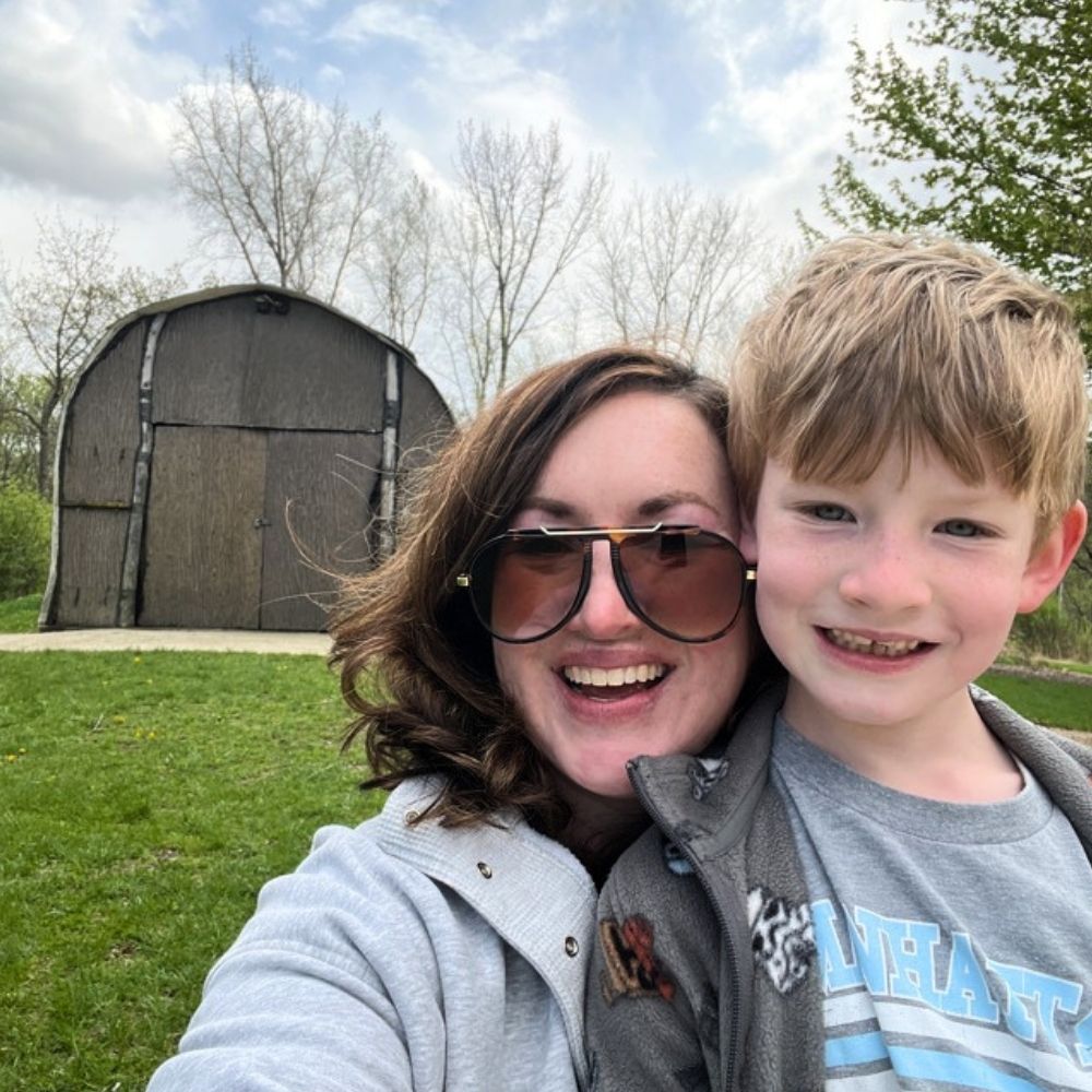 A smiling woman wearing large sunglasses poses with a young child in front of a historic longhouse structure in a grassy park setting.