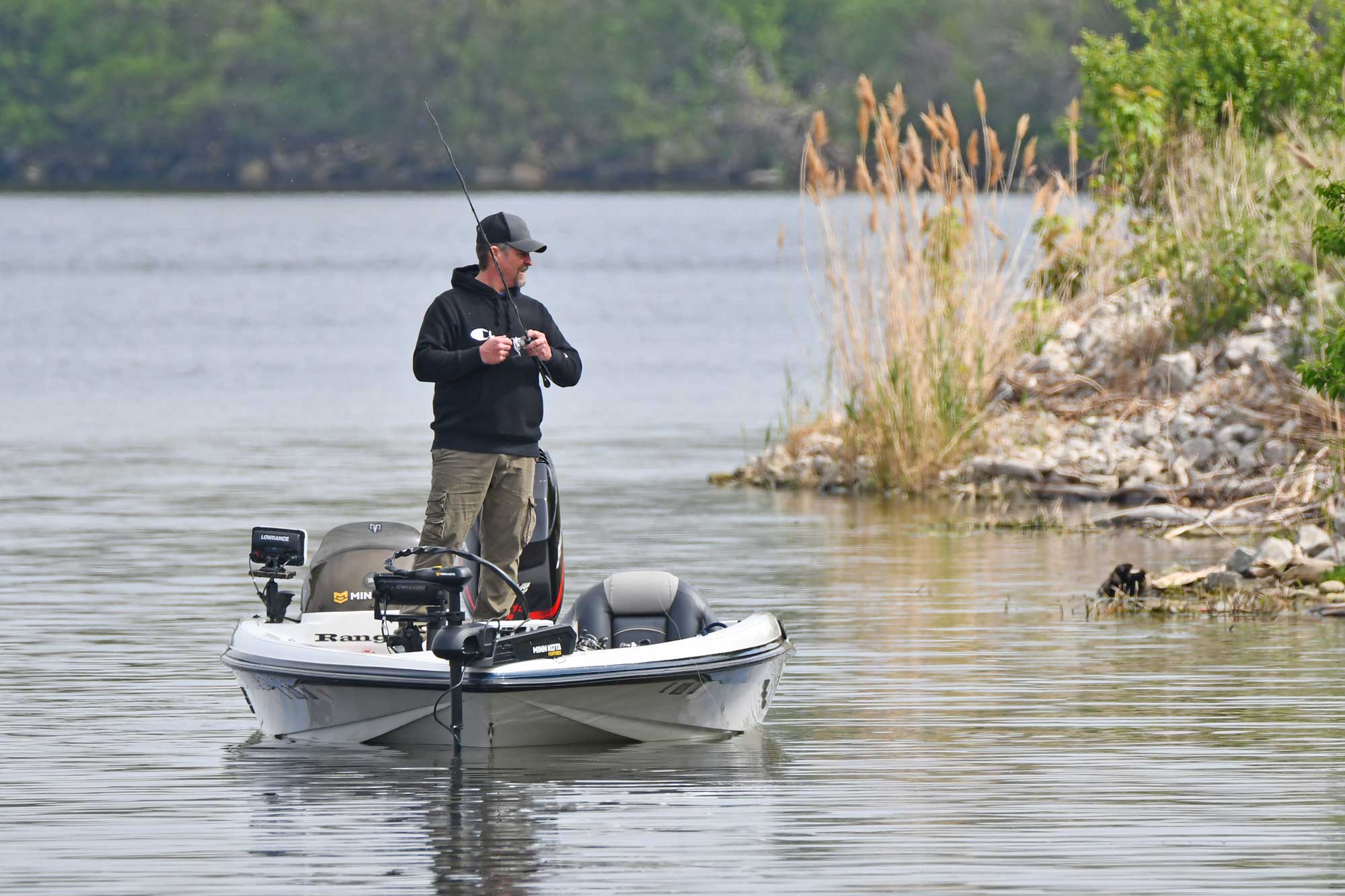 Person standing up in a boat while fishing with trees in the background.