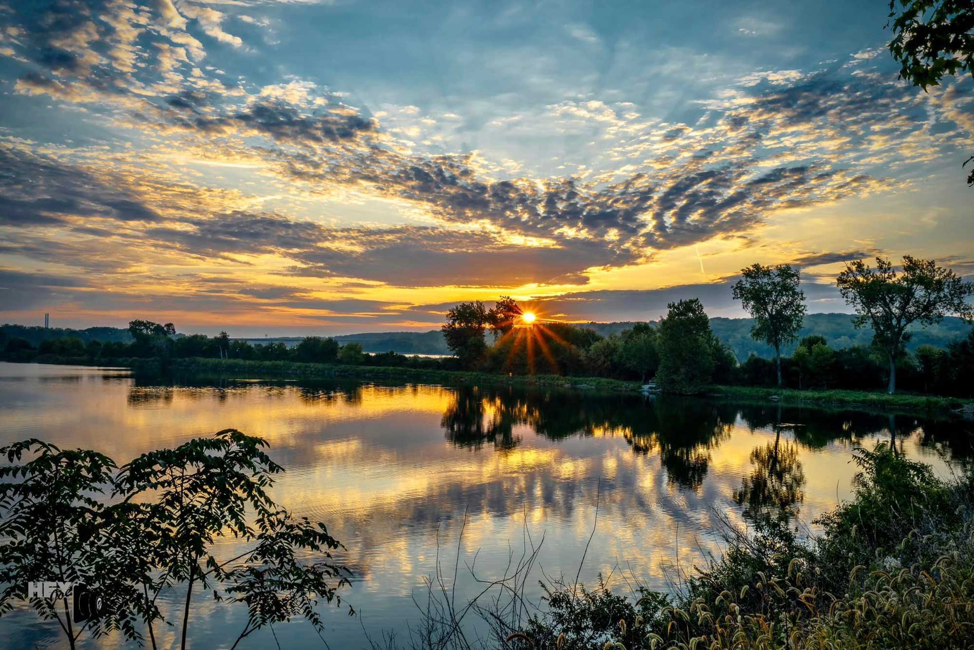 Lake with paved trail, trees, and the rising sun in the background.