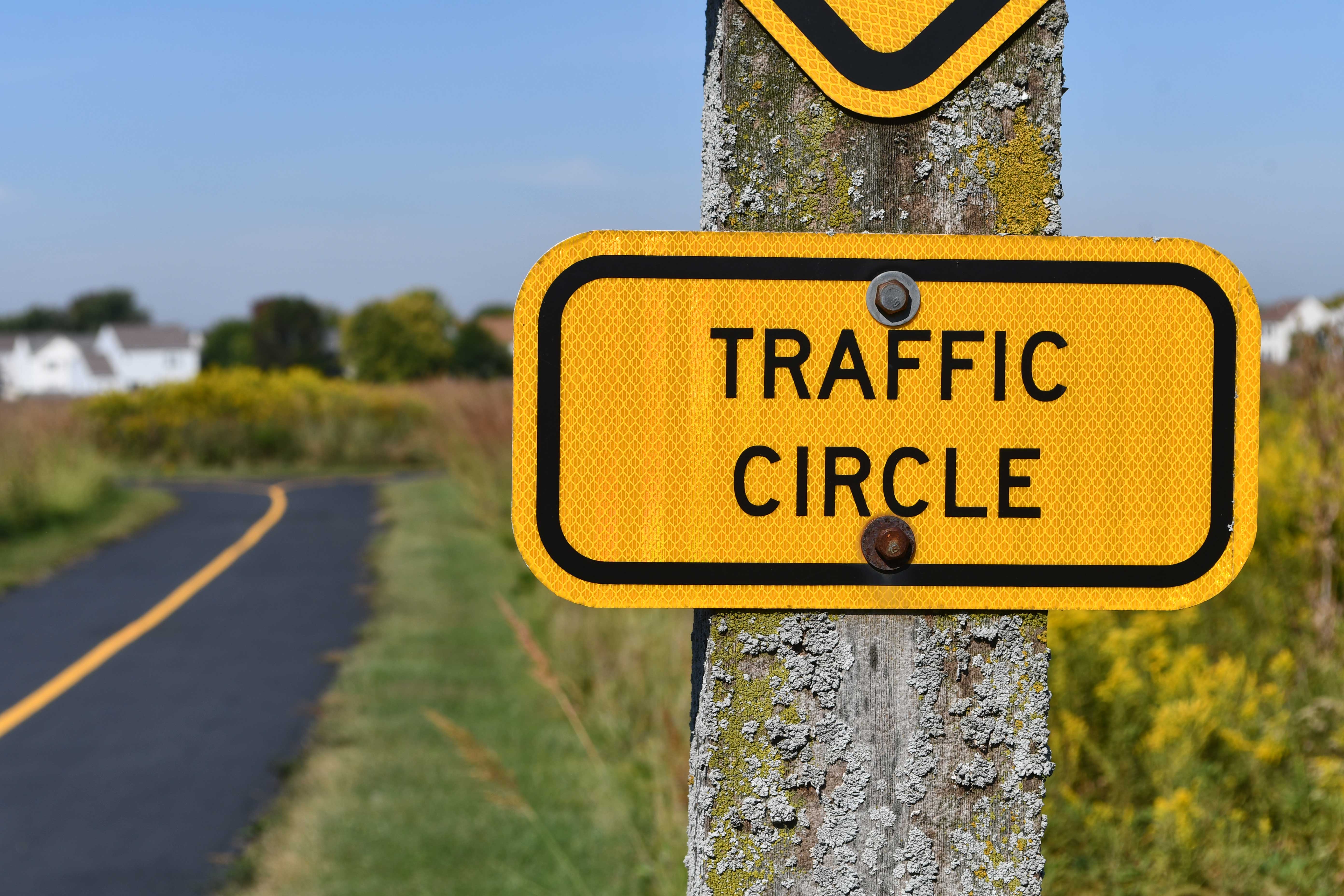 Close-up of a yellow “Traffic Circle” sign mounted on a weathered wooden post covered in lichen, with a paved trail curving through prairie grasses and homes visible in the distance.