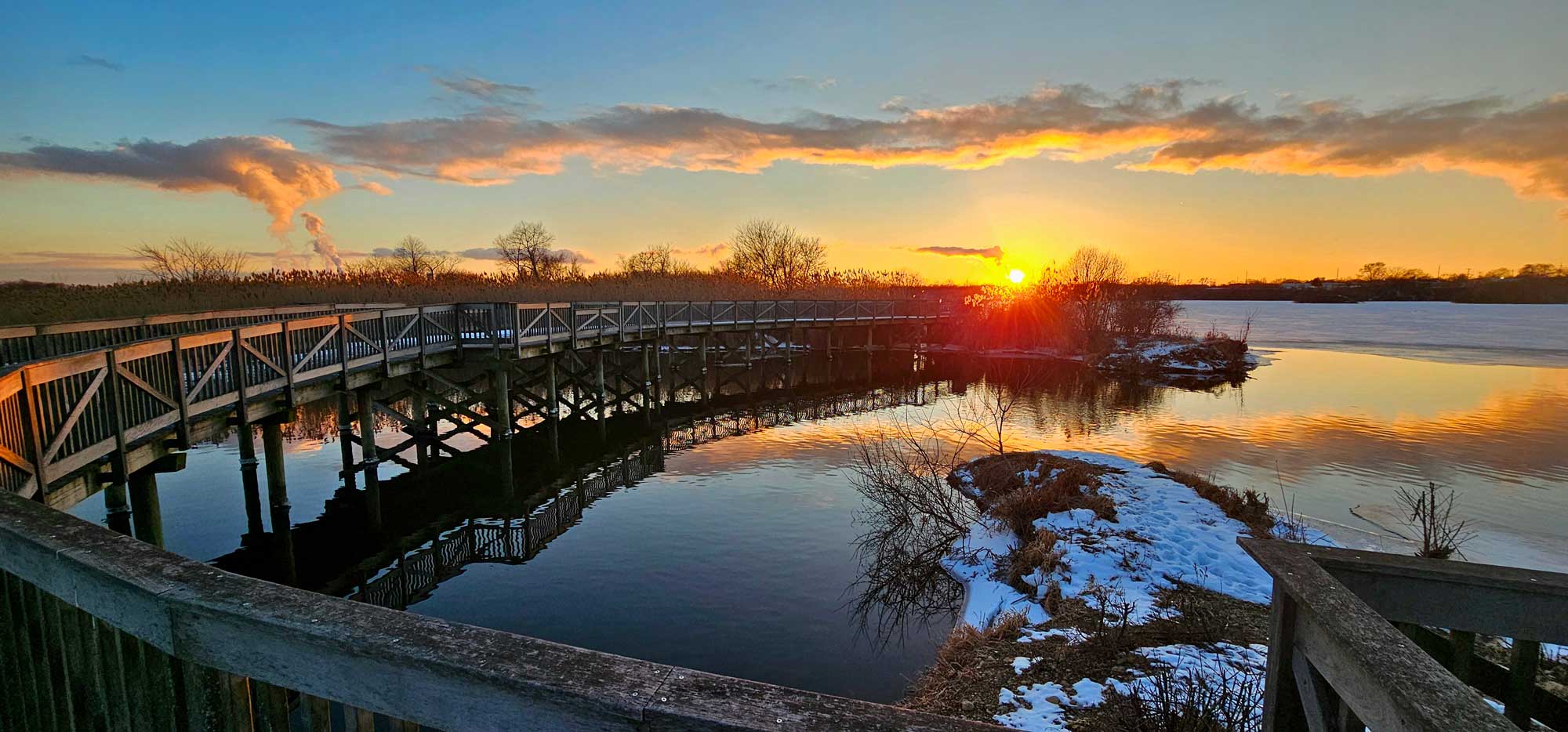 Boardwalk over water with trees and the setting sun in the background.