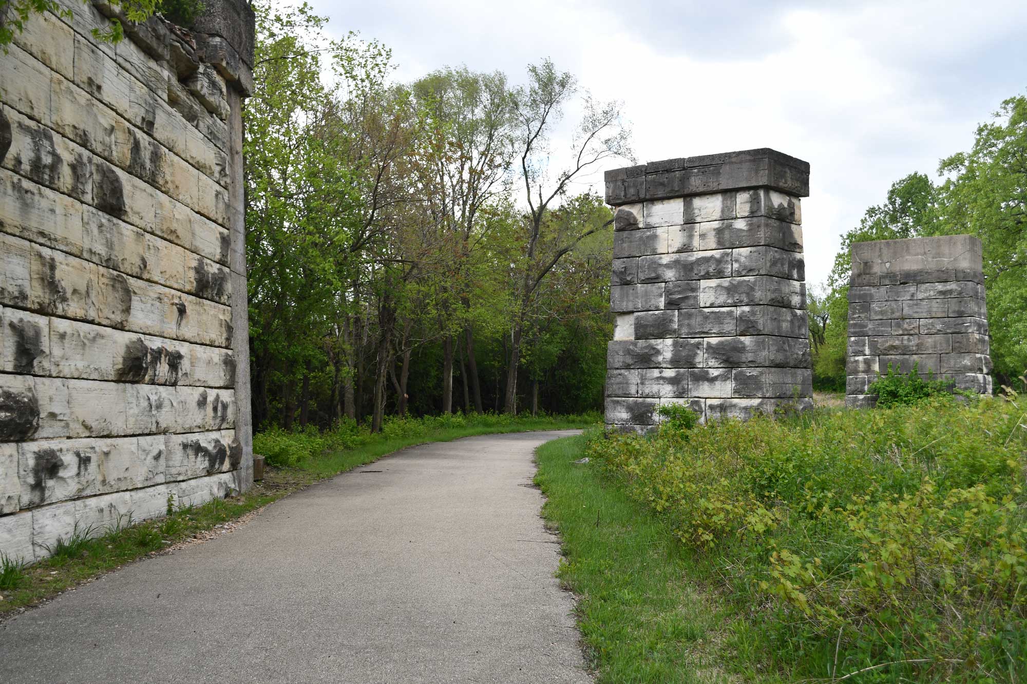 Still standing ruins along paved trail with grasses, vegetation, and trees nearby.