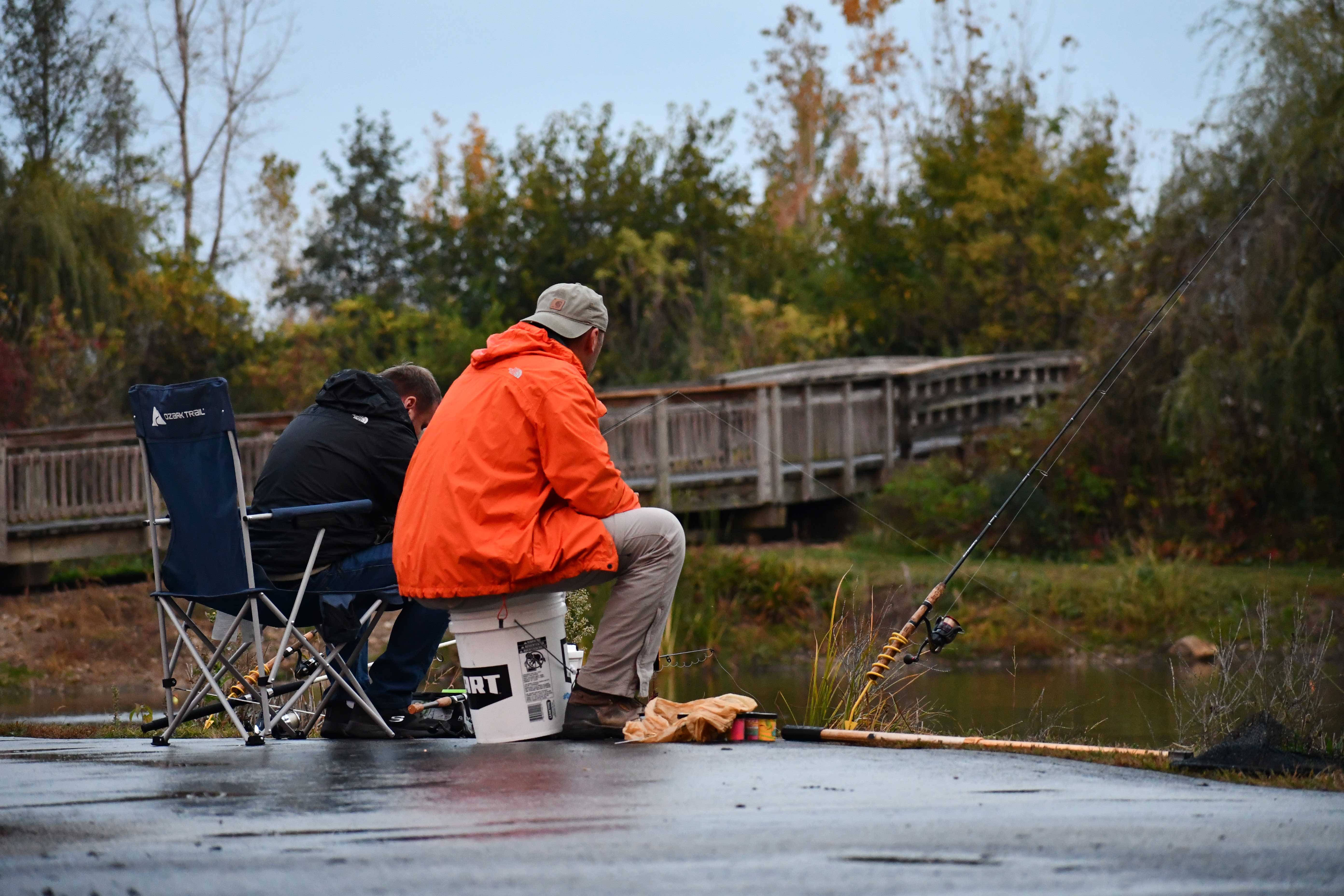 Two people sit near a pond on a damp day, fishing together. One person wears an orange rain jacket and sits on a bucket, while the other wears a dark jacket and sits in a folding chair.